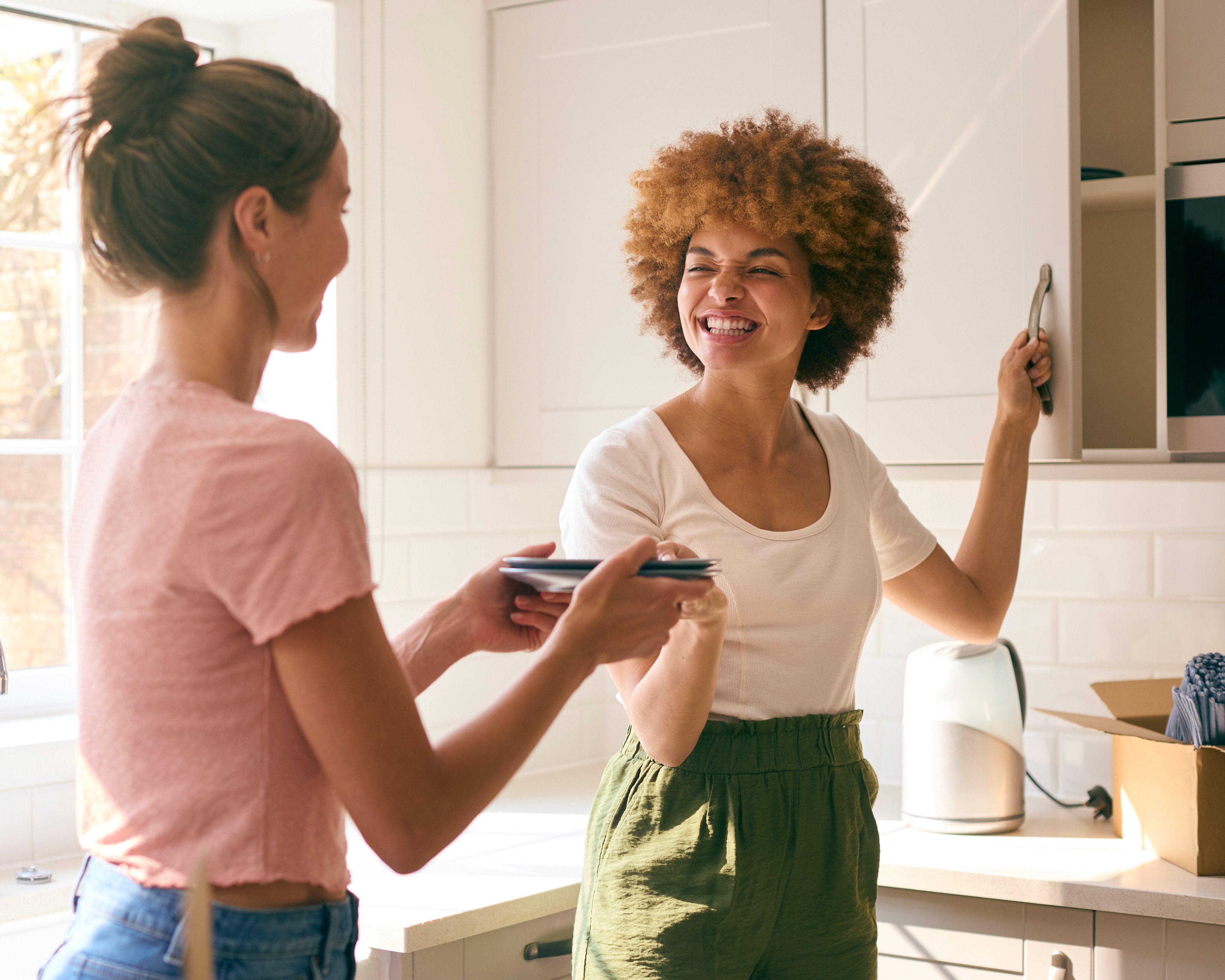 Two women are unpacking plates from a moving box into a cupboard and smiling at one another.