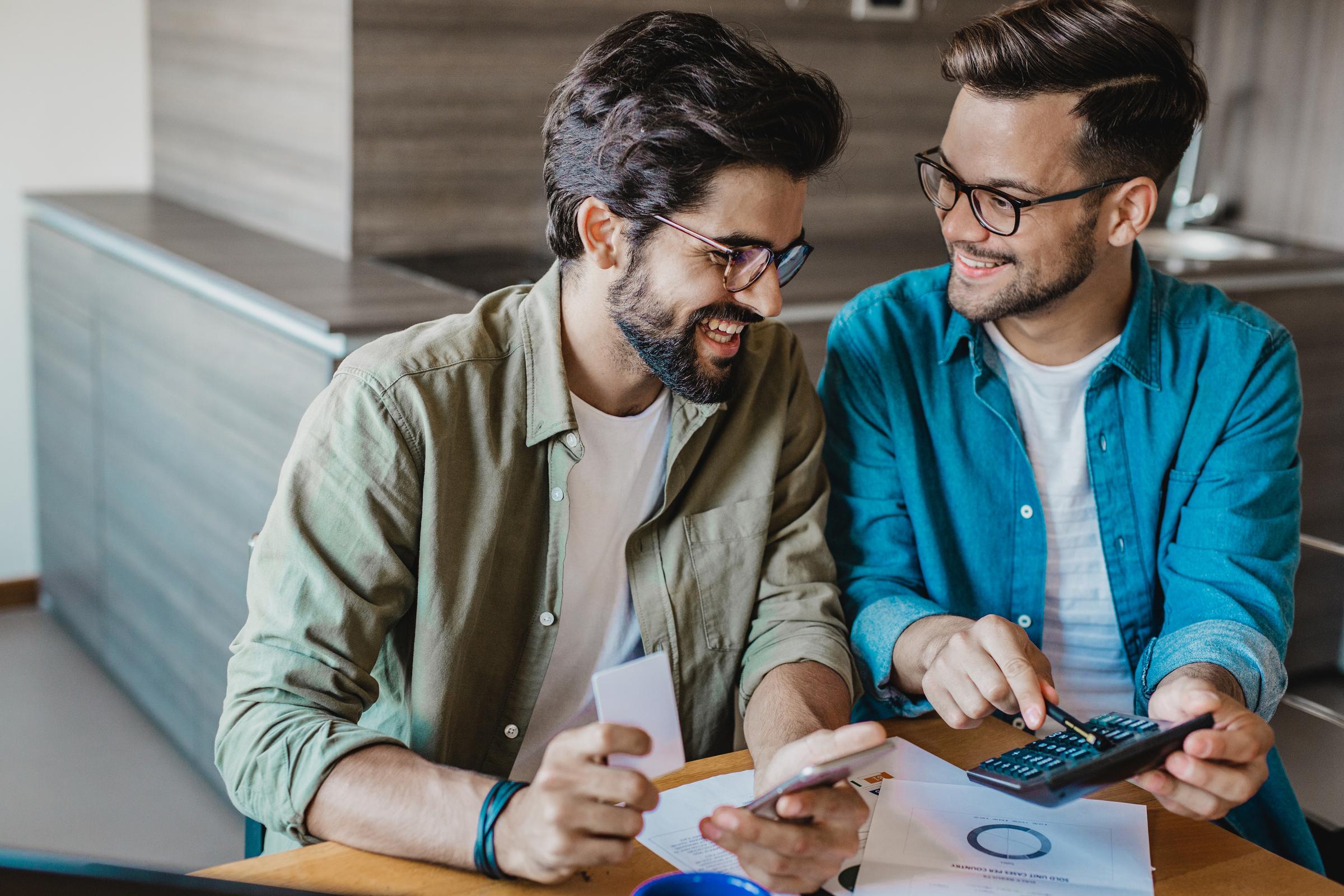 Couple at their dining table. One man holds his phone and a card and the other man holds calculator