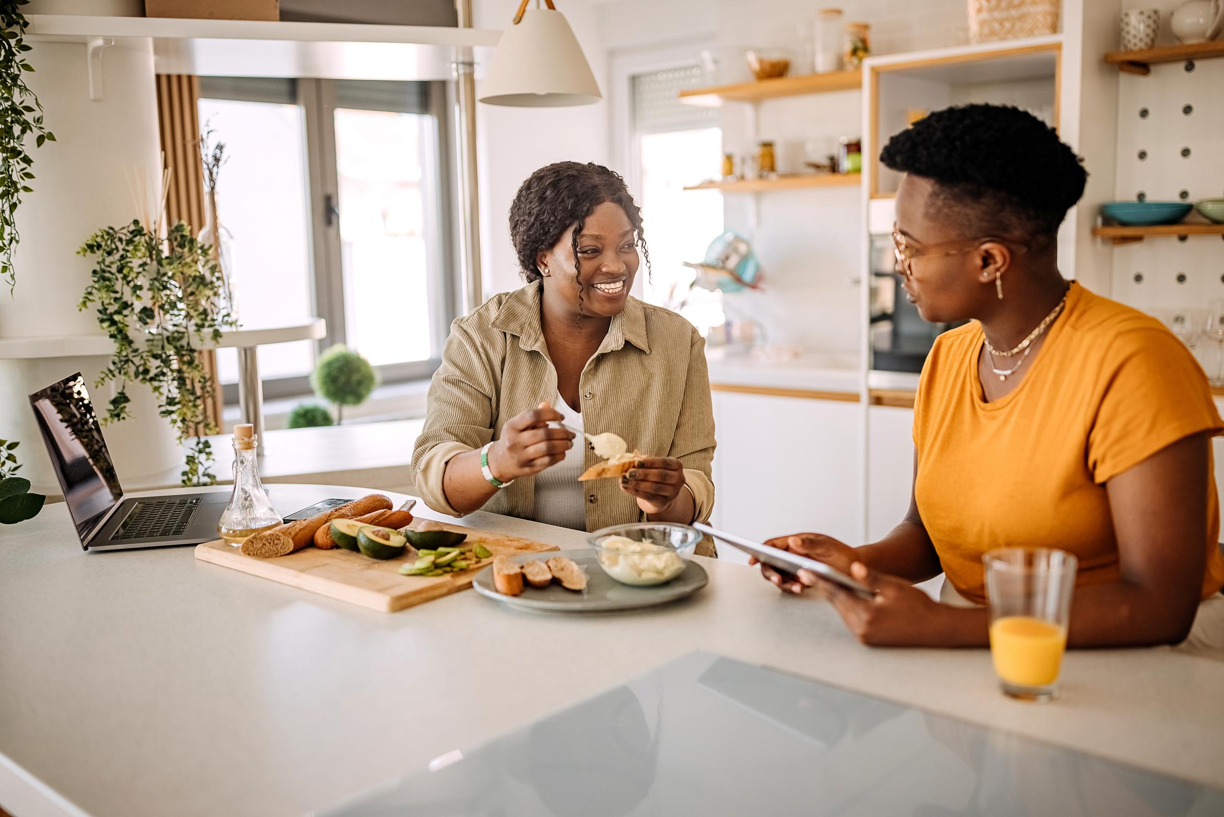 Two women sit at a dining table. One is making lunch and another holds a touchpad.