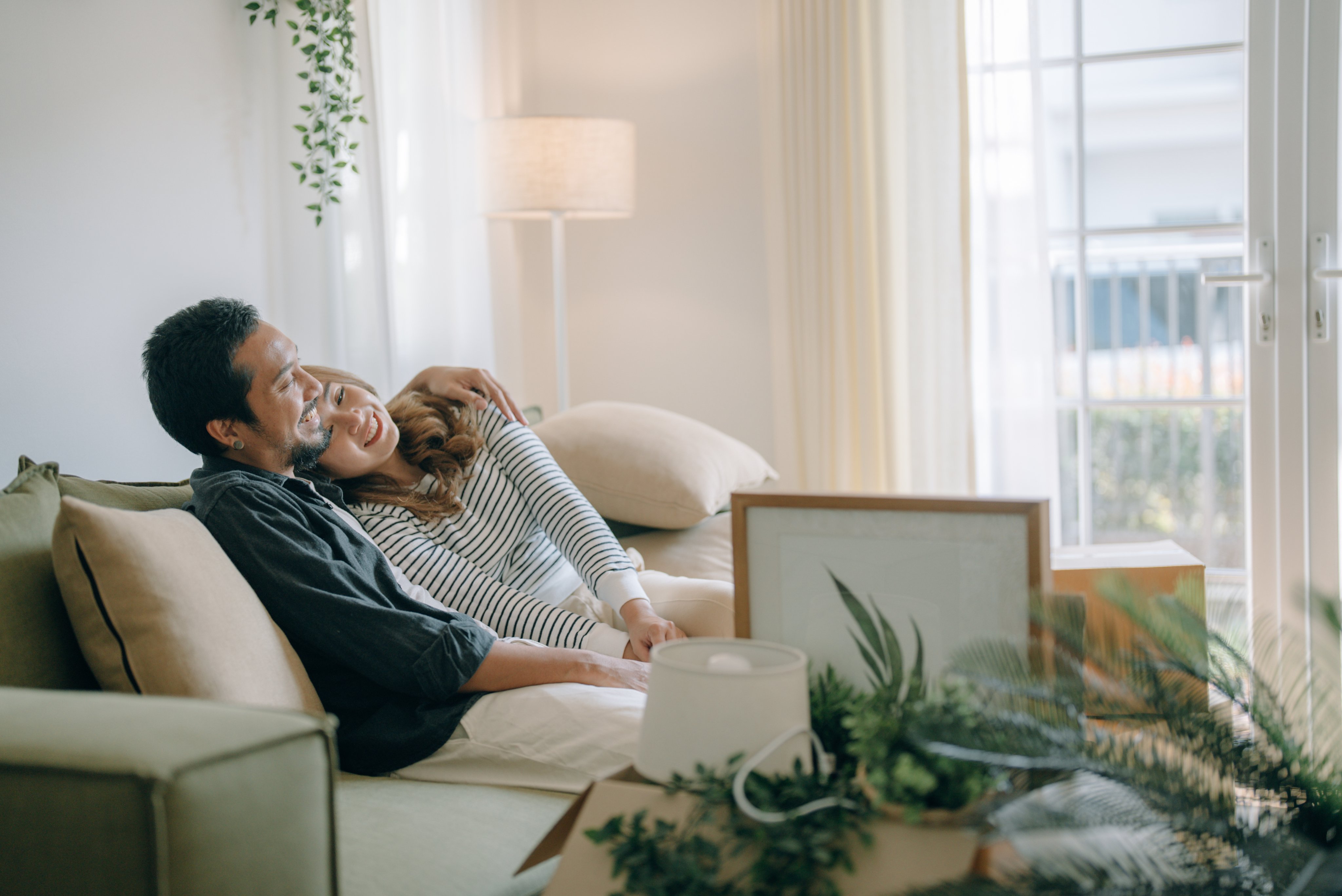A man and woman embrace on a sofa, surrounded by moving boxes, containing plants, frames and a lamp.