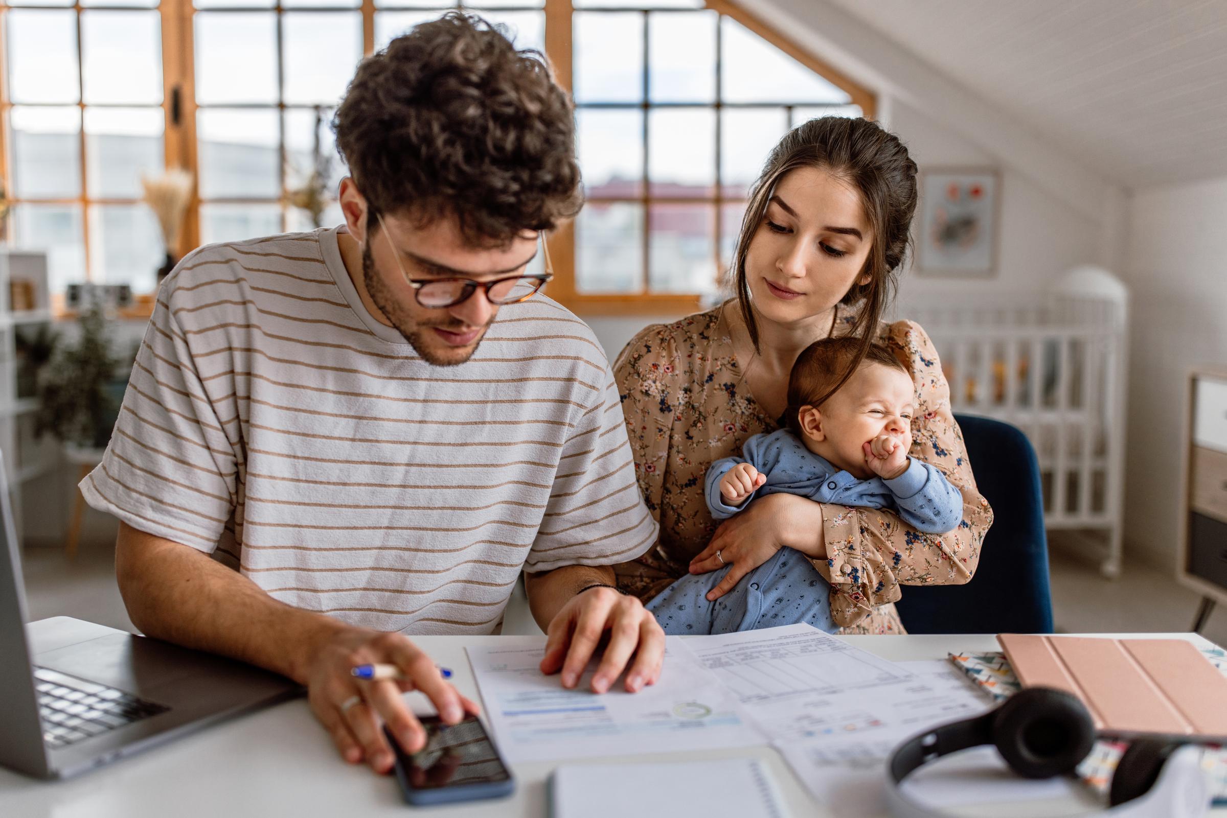 A couple look at a piece of paper showing finances. The Woman holds baby and the man holds phone.