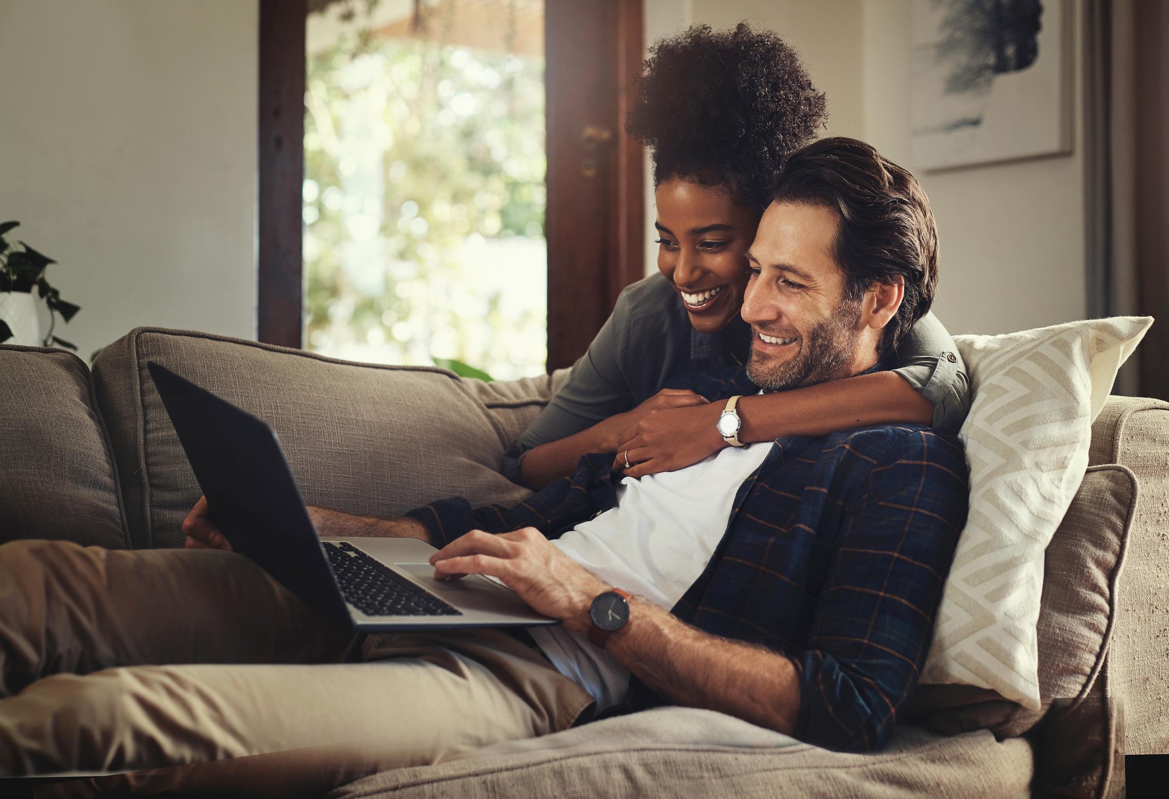 Couple on their sofa smiling looking at a laptop. The woman hugs the man.