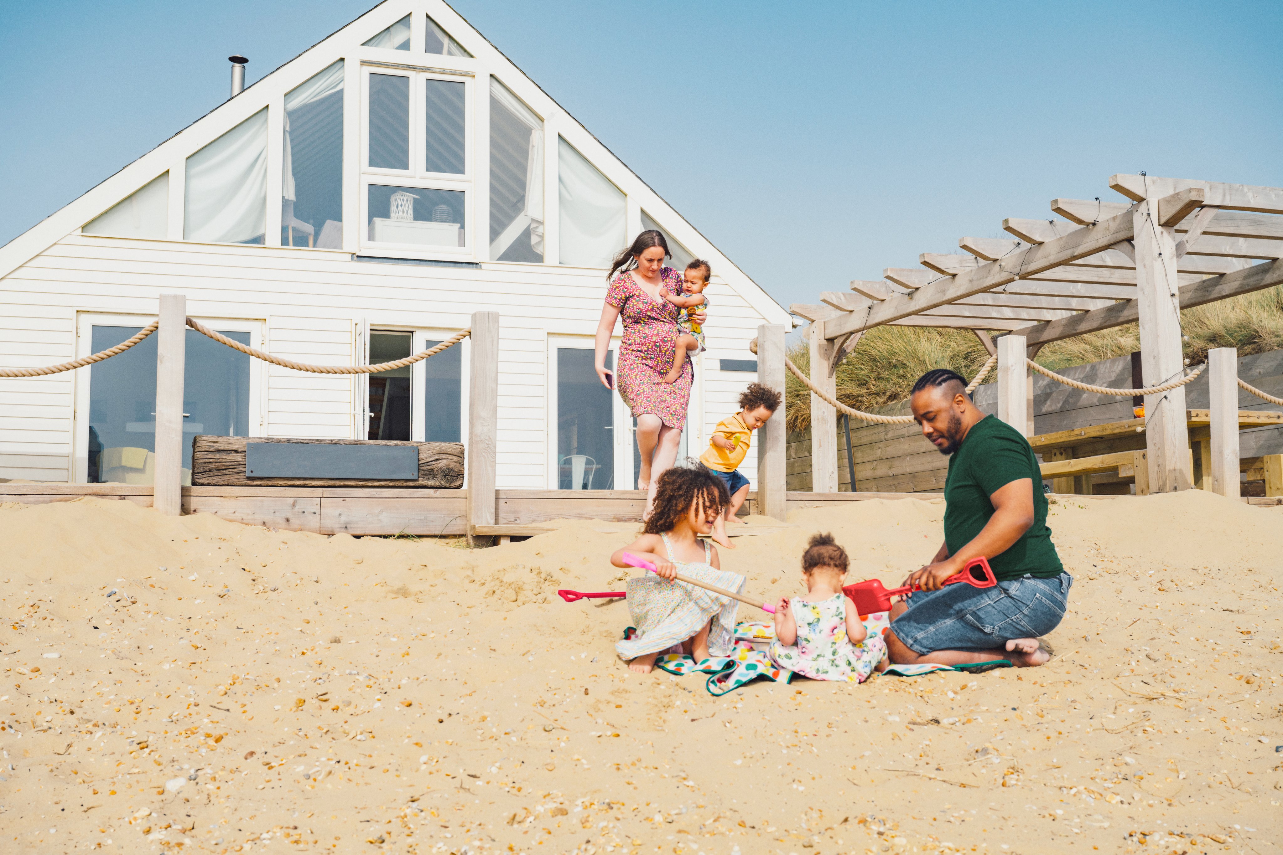 A family are at a beach house. Dad is building sandcastles with his 2 children. Mum carries the baby and a young child is running.