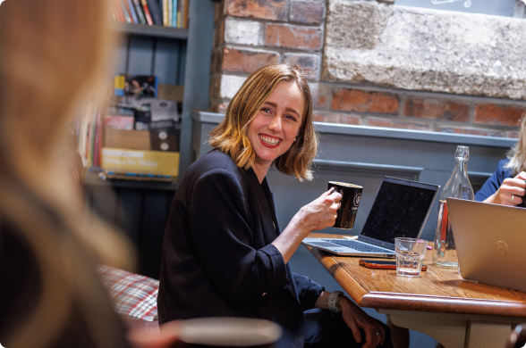 A woman with a mug smiling while working at laptop in a coffee shop