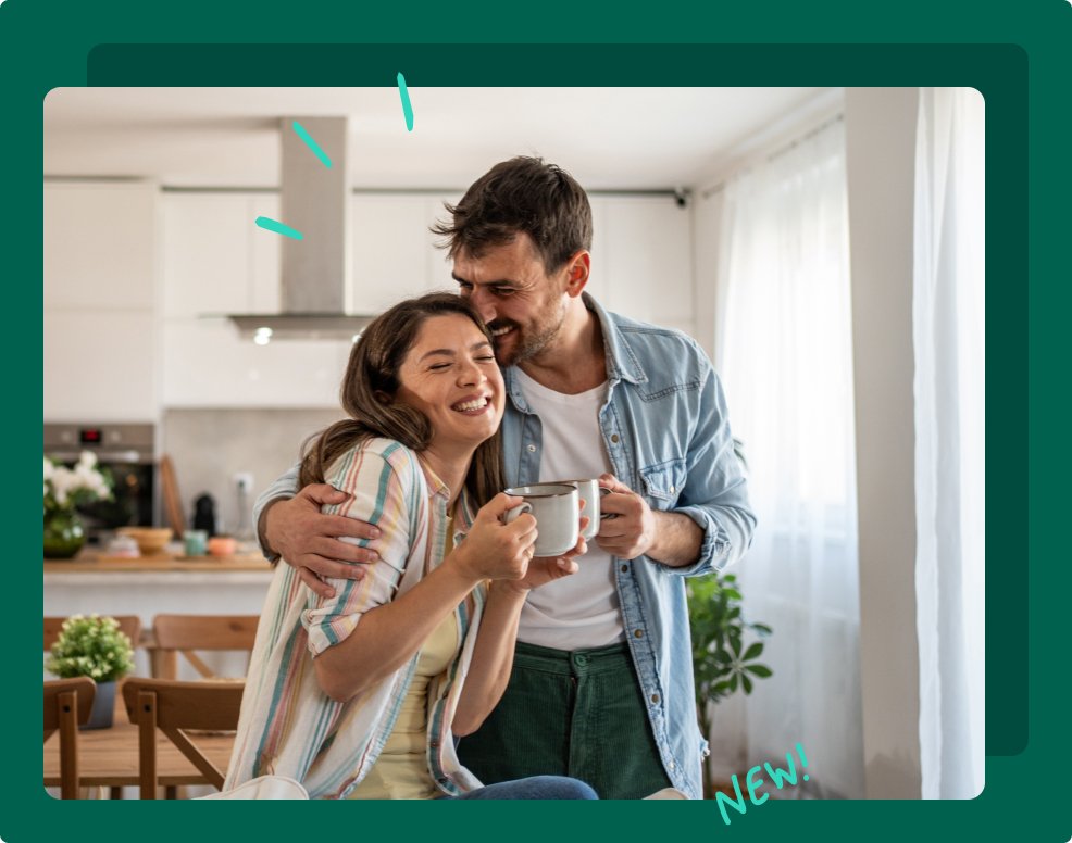 Smiling couple hugging in a kitchen while holding coffee mugs.