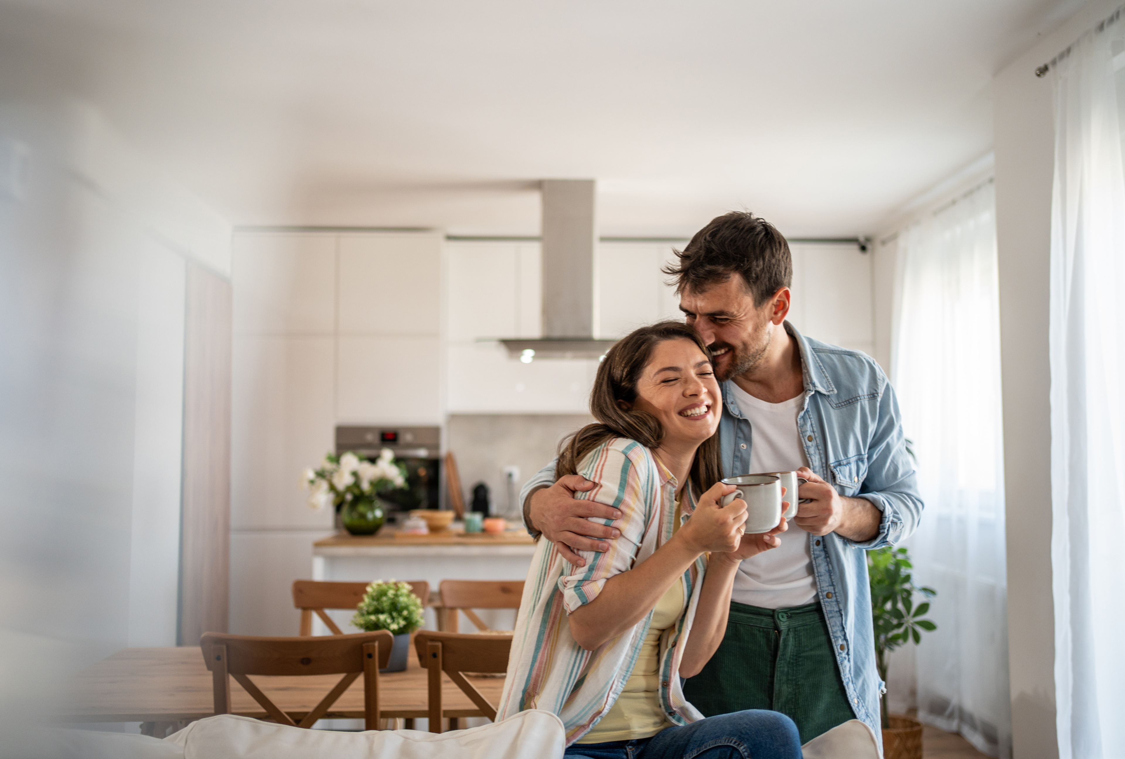 Smiling couple hugging in a kitchen while holding coffee mugs.