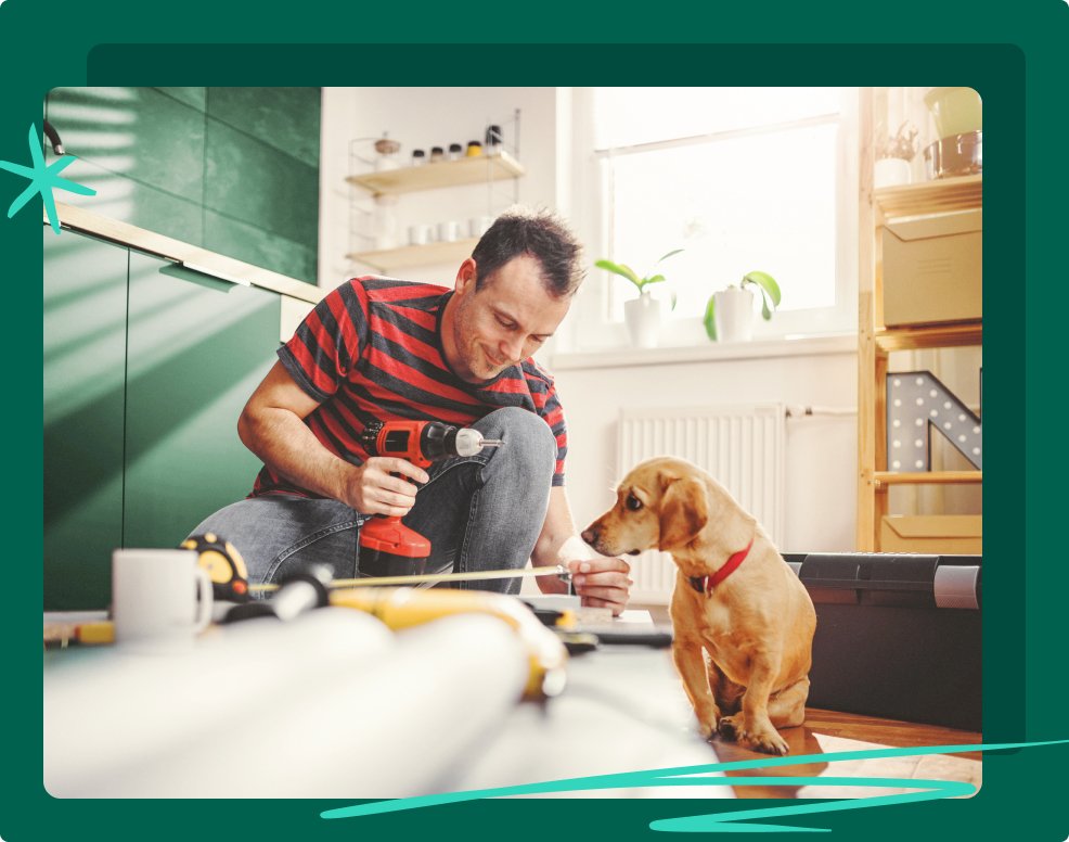 A man is holding a drill and doing some DIY in his kitchen. His dog is sat watching him.