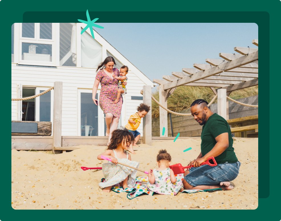 A family are at a beach house. Dad is building sandcastles with his 2 children. Mum carries the baby and a young child is running.