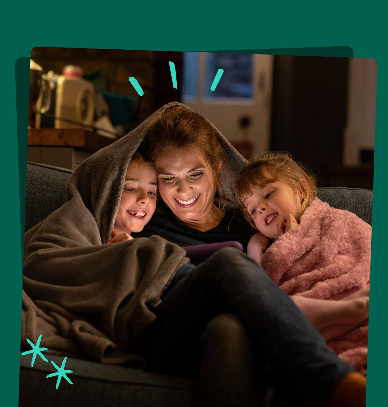 A shot of two young girls with their mother, sitting comfortably with a blanket over their heads on a sofa in the living room of their home