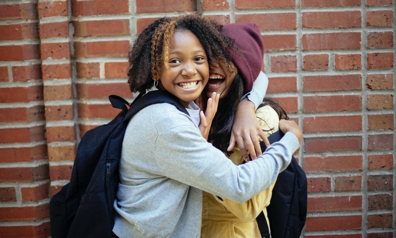 Two children wearing backpacks hugging and smiling at the camera.