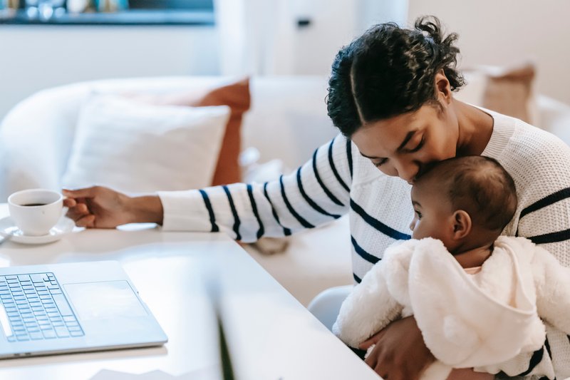 Woman kissing a baby's head as she holds them in one hand while the other holds a coffee cup, that sits on a table next to a laptop.