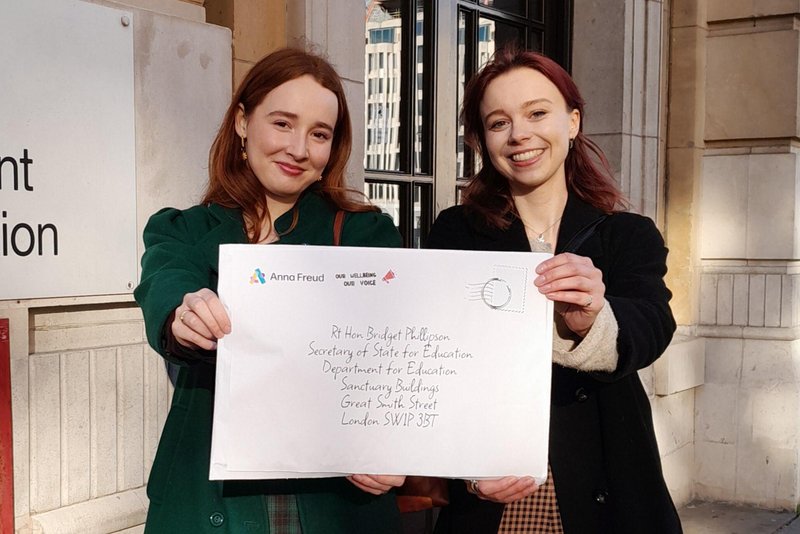 Grace and Emily outside Westminster holding up a letter to support the Our Wellbeing Our Voice campaign.