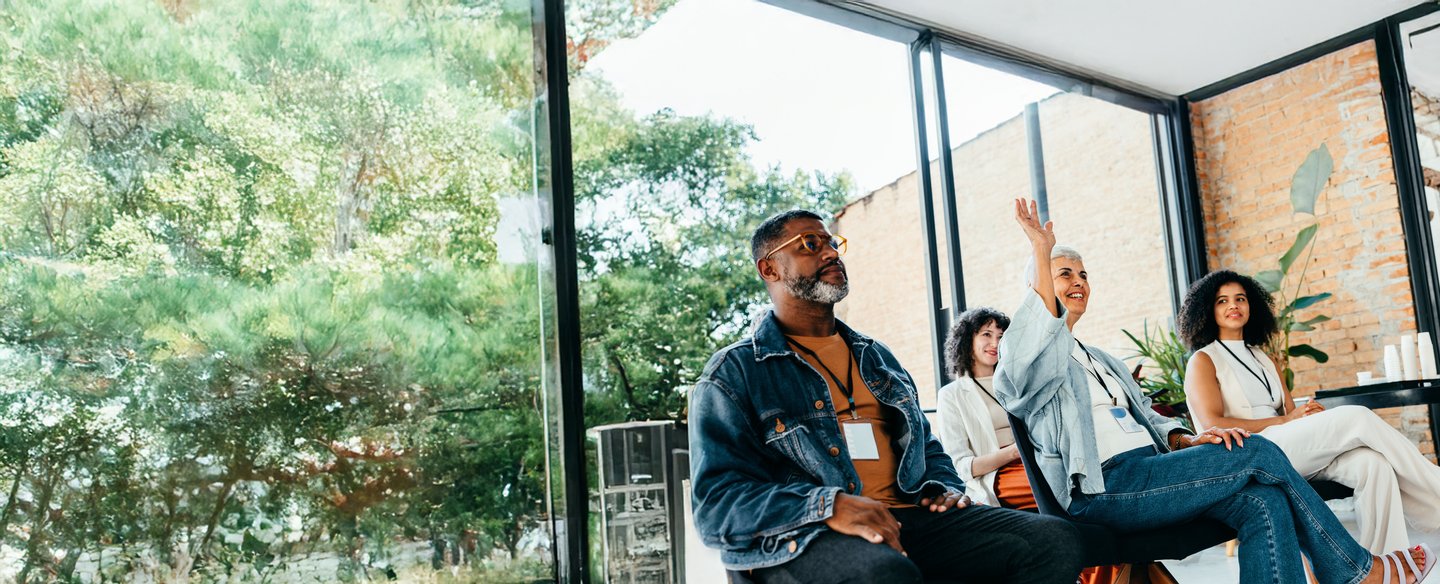 Five professionals wearing lanyards at a conference. A woman in the foreground has her hand raised, while the person to her left looks at her and smiles.