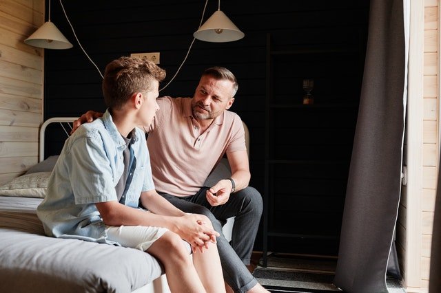 A man faces the camera while placing a hand on a young person’s back as they sit together on a bed.