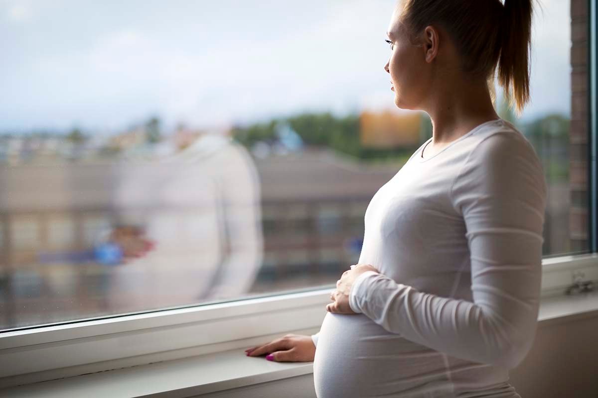 Pregnant woman looks out large window with hand on belly