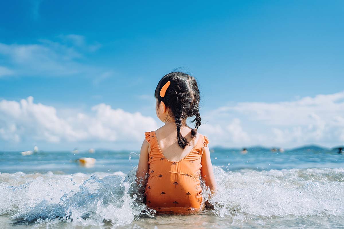 Little girl in bright orange swimsuit sits at edge of ocean