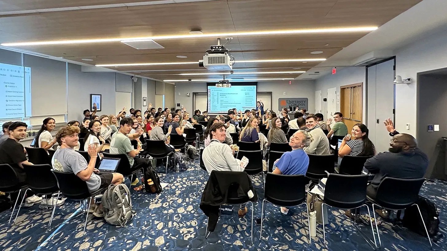 Students in the 2026 Introduction to Clinical Research course turn in their seats and wave at the camera in a UVA classroom.