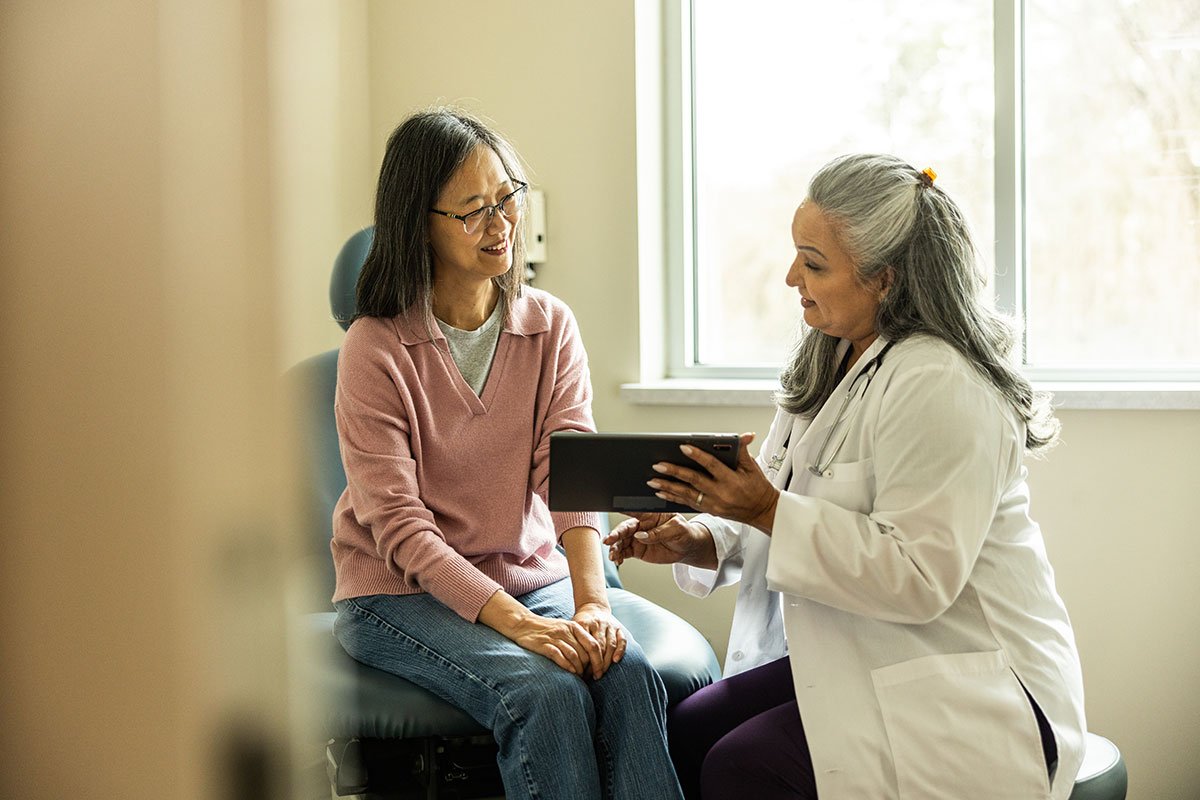 Two people talking in a clinical setting, one holding an iPad and wearing a white coat.