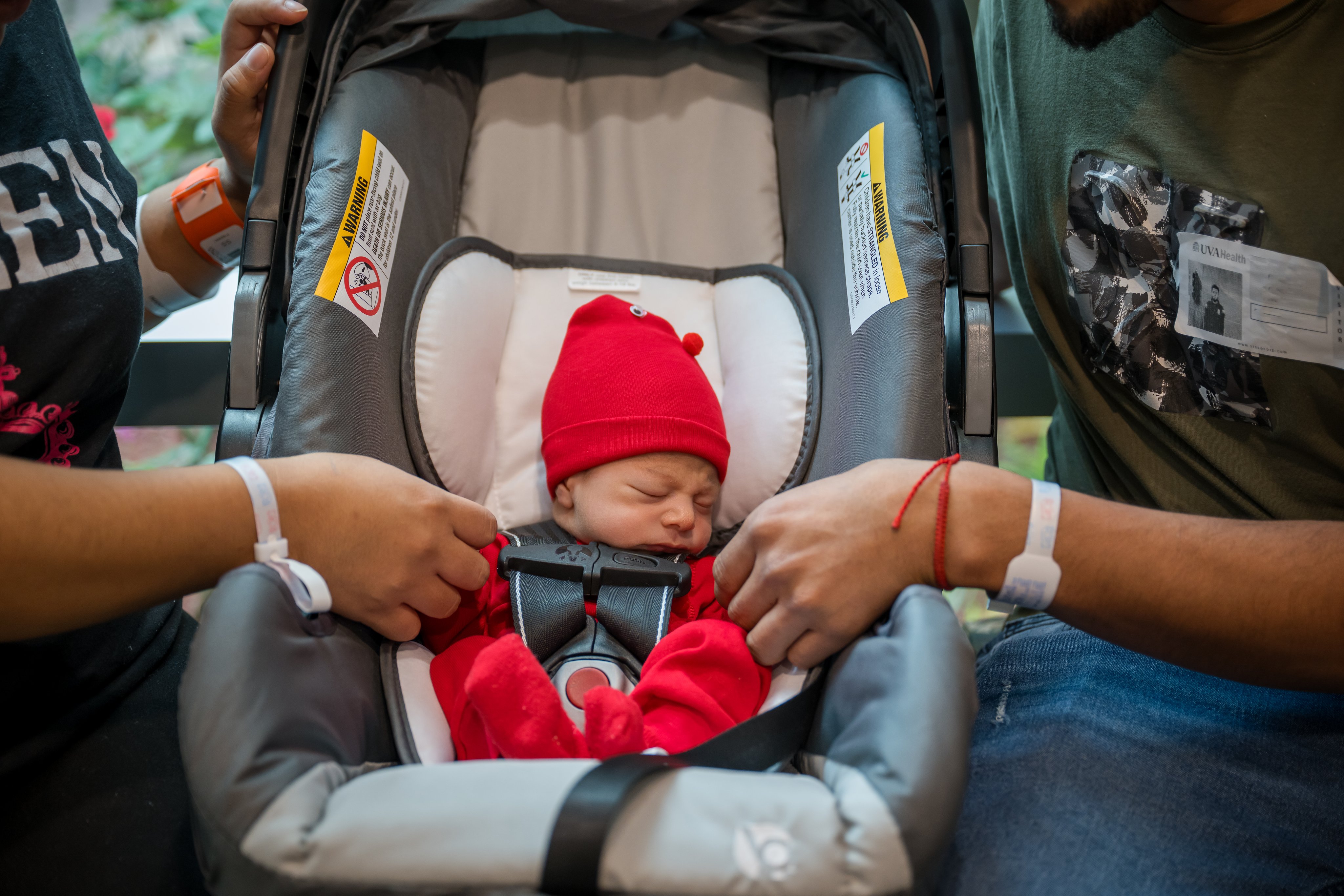 A baby sleeps in a carseat while holding both parents hands 