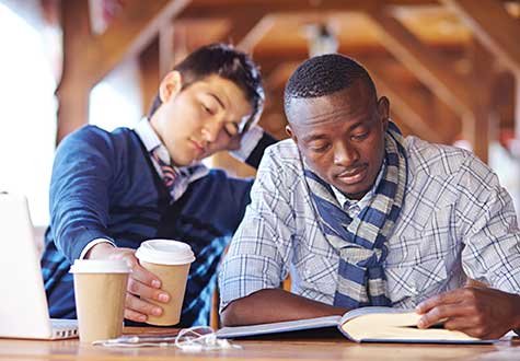 Two college-aged boys drink coffee while looking over a textbook