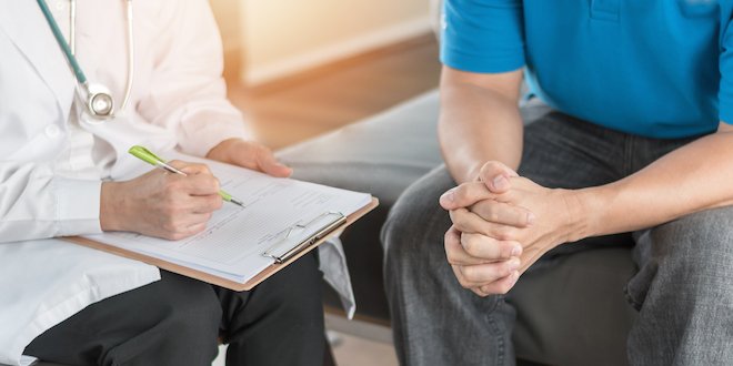 Doctor takes notes while man folds hands in front of him. 