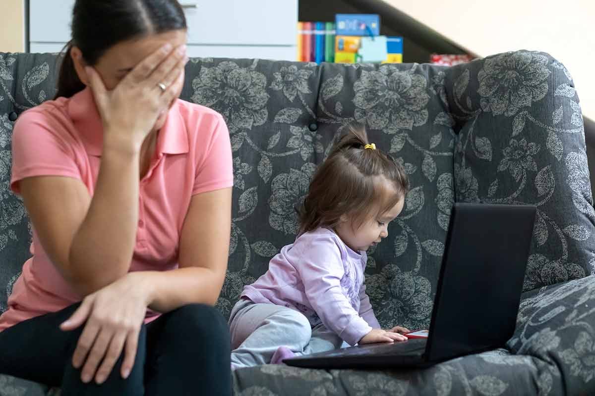 overwhelmed mom rests head in hands while toddler bangs on laptop