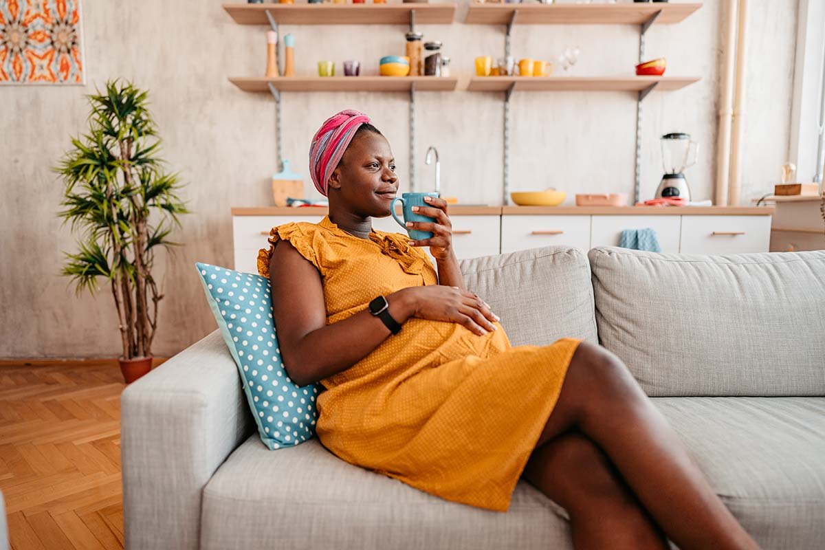 Pregnant woman sips tea and sits on couch with smile, with one hand on her belly
