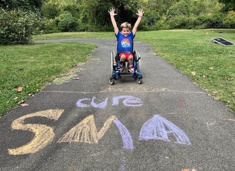 young boy in wheelchair outside with "cure SMA" written in sidewalk chalk in front of him