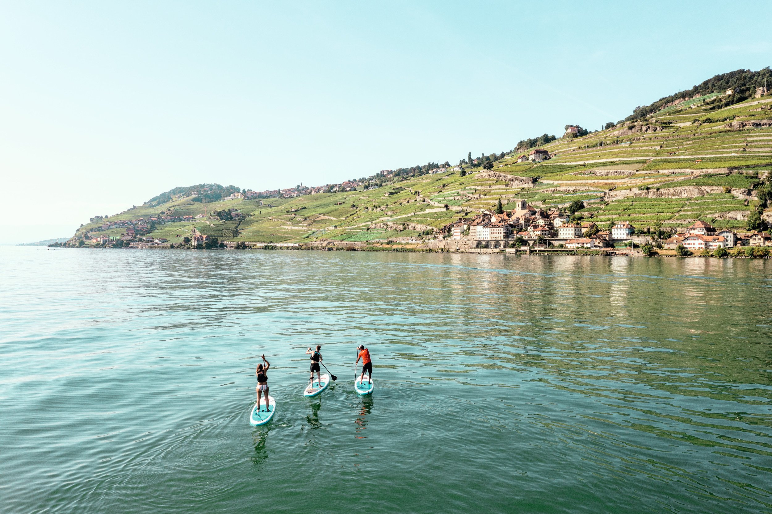 Three people stand-up paddleboarding on Lake Geneva with the Lavaux vineyards in the background and a blue, cloudless sky. Drone image from above.