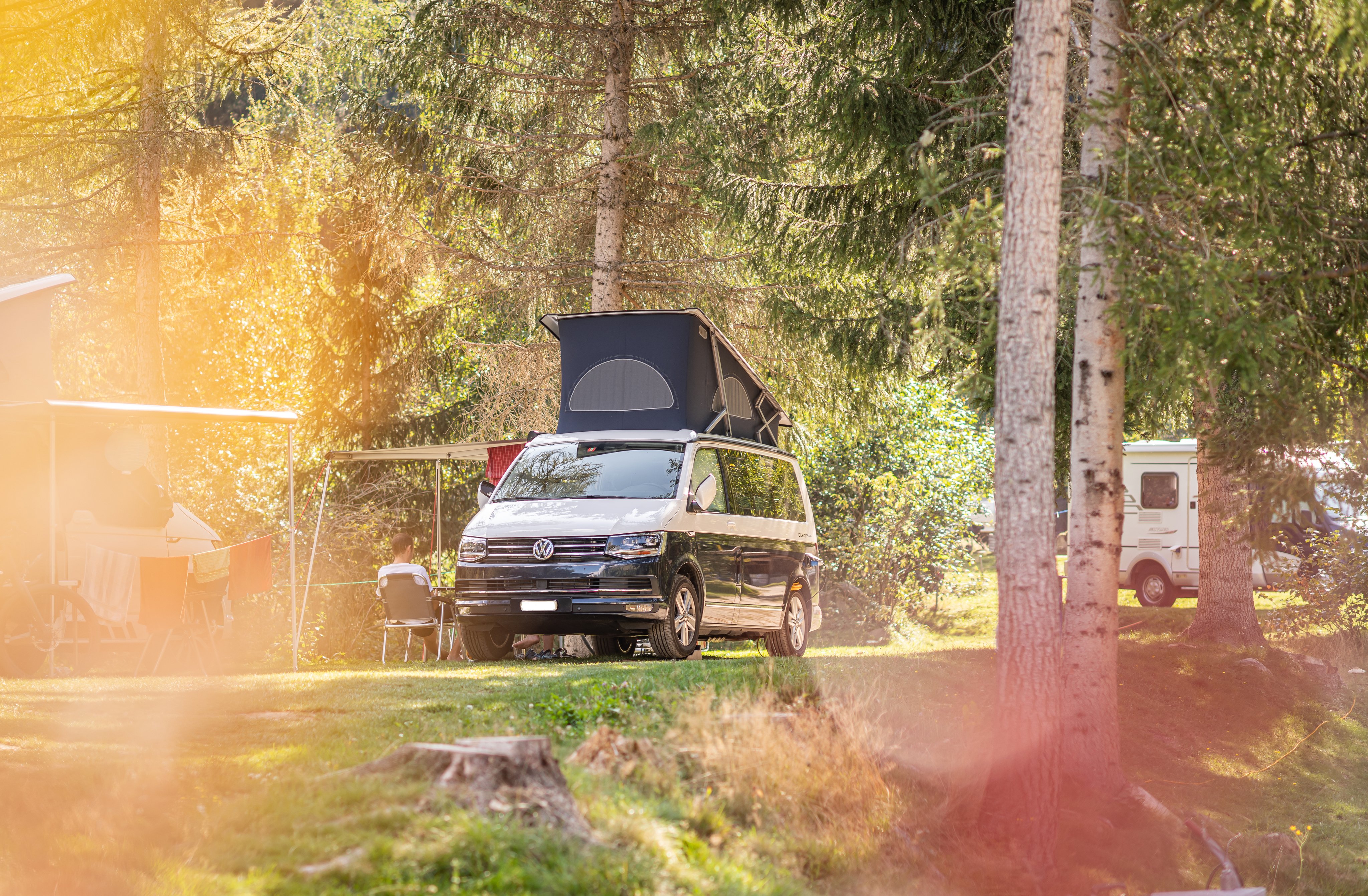 Ein VW-Camper mit hochgestelltem Dach steht unter Bäumen auf einem Campingplatz.