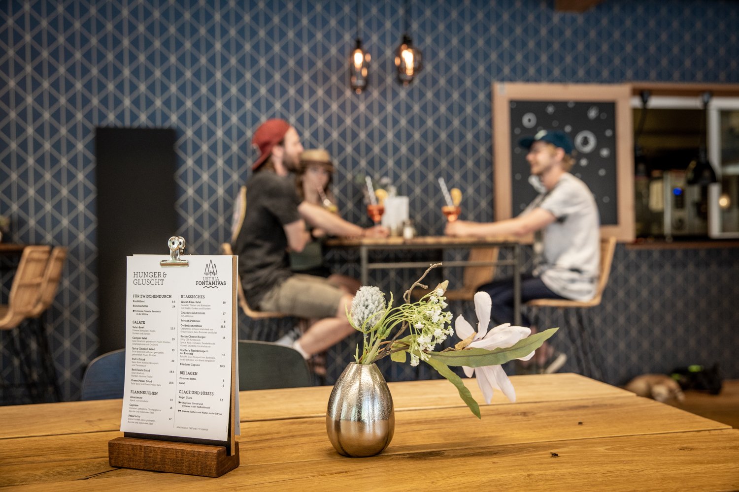 Un restaurant avec un menu au premier plan et trois personnes à une table haute derrière.