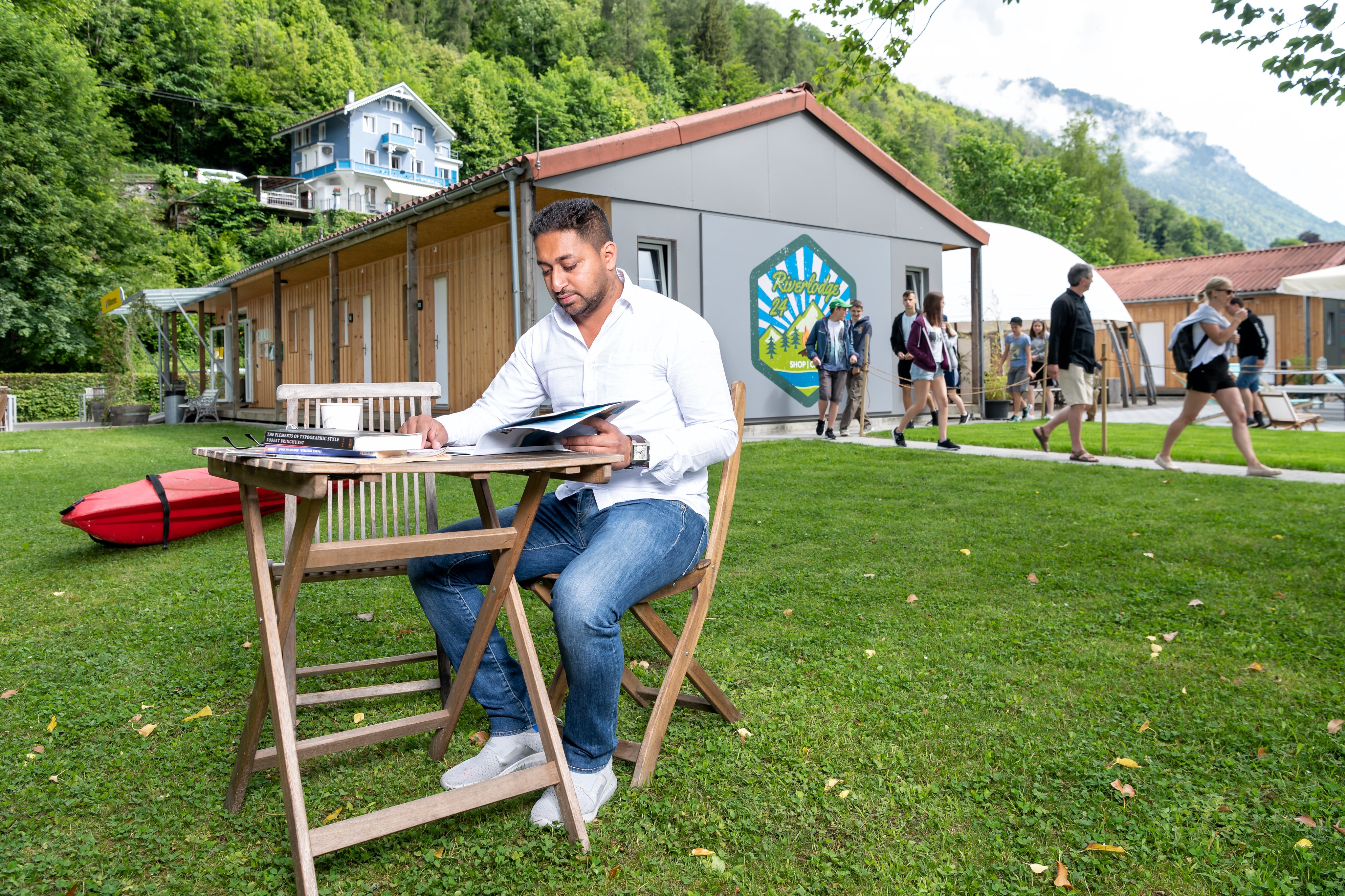 Ein Mann sitzt vor einem Gebäude an einem Tisch und liest in einem Magazin.