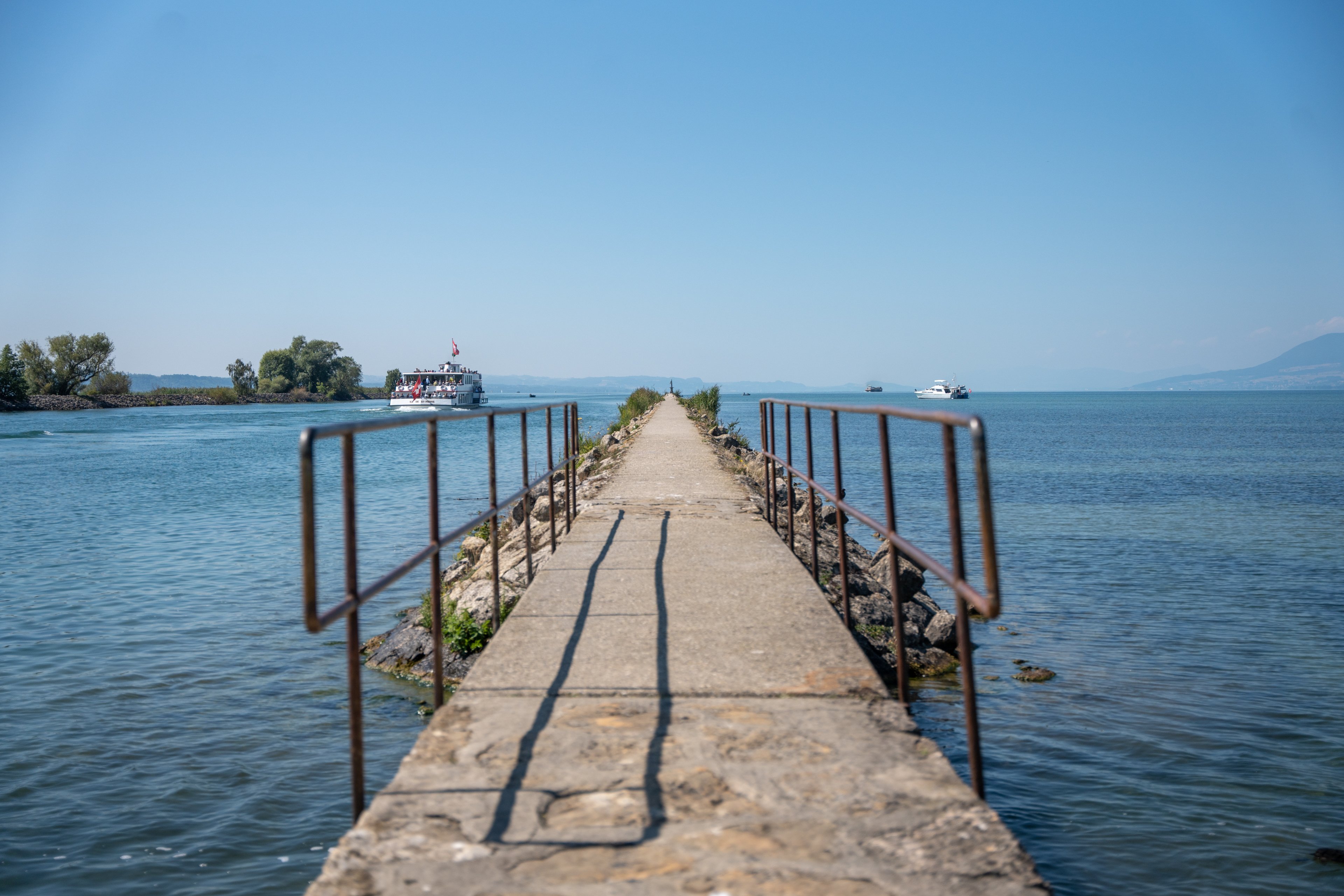 Ein Steg in den See. Ein Schiff und ein Boot im Hintergrund.
