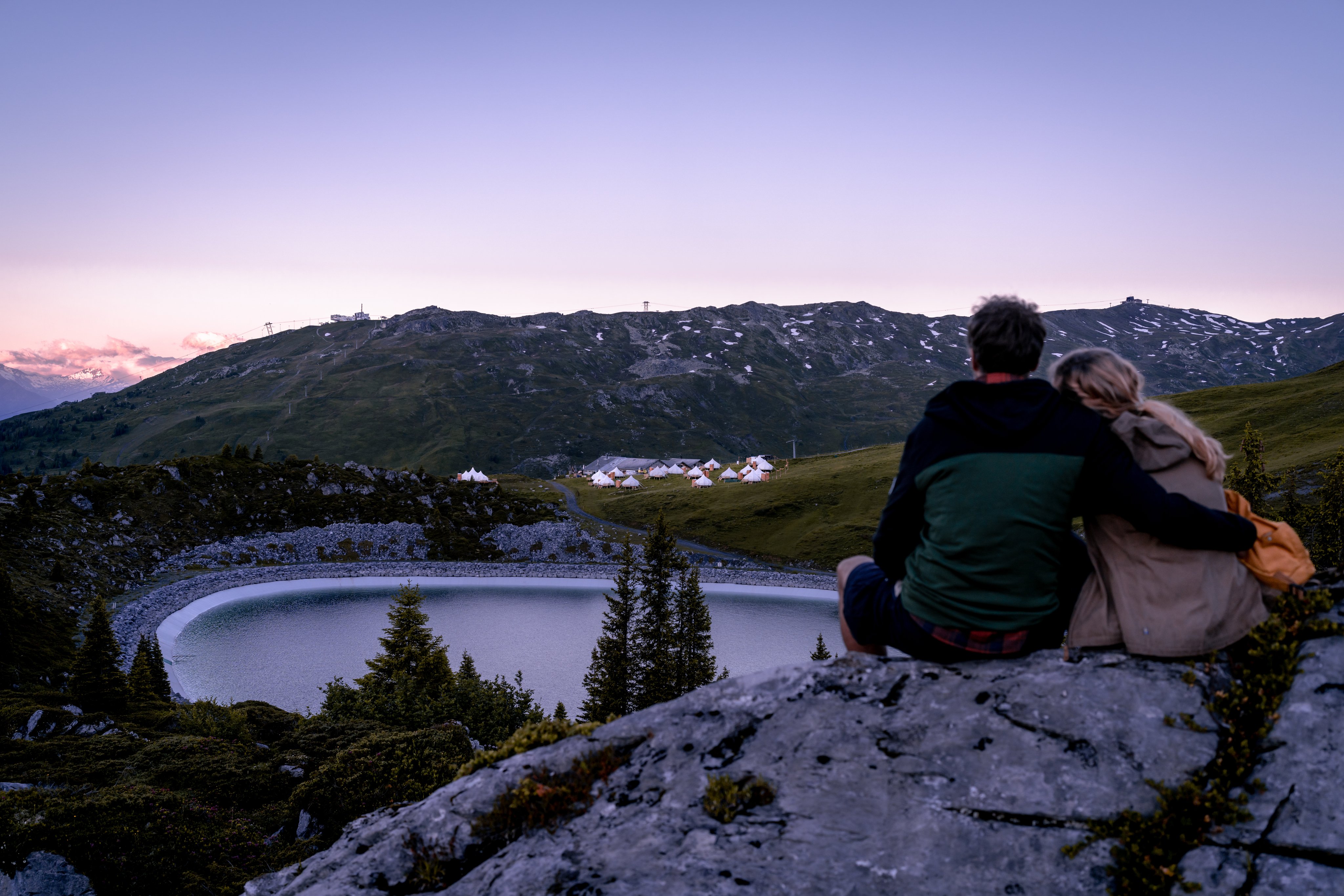 Un couple est assis sur un rocher avec vue sur le TCS Pop-Up Glamping Laax, le lac artificiel et les montagnes environnantes.