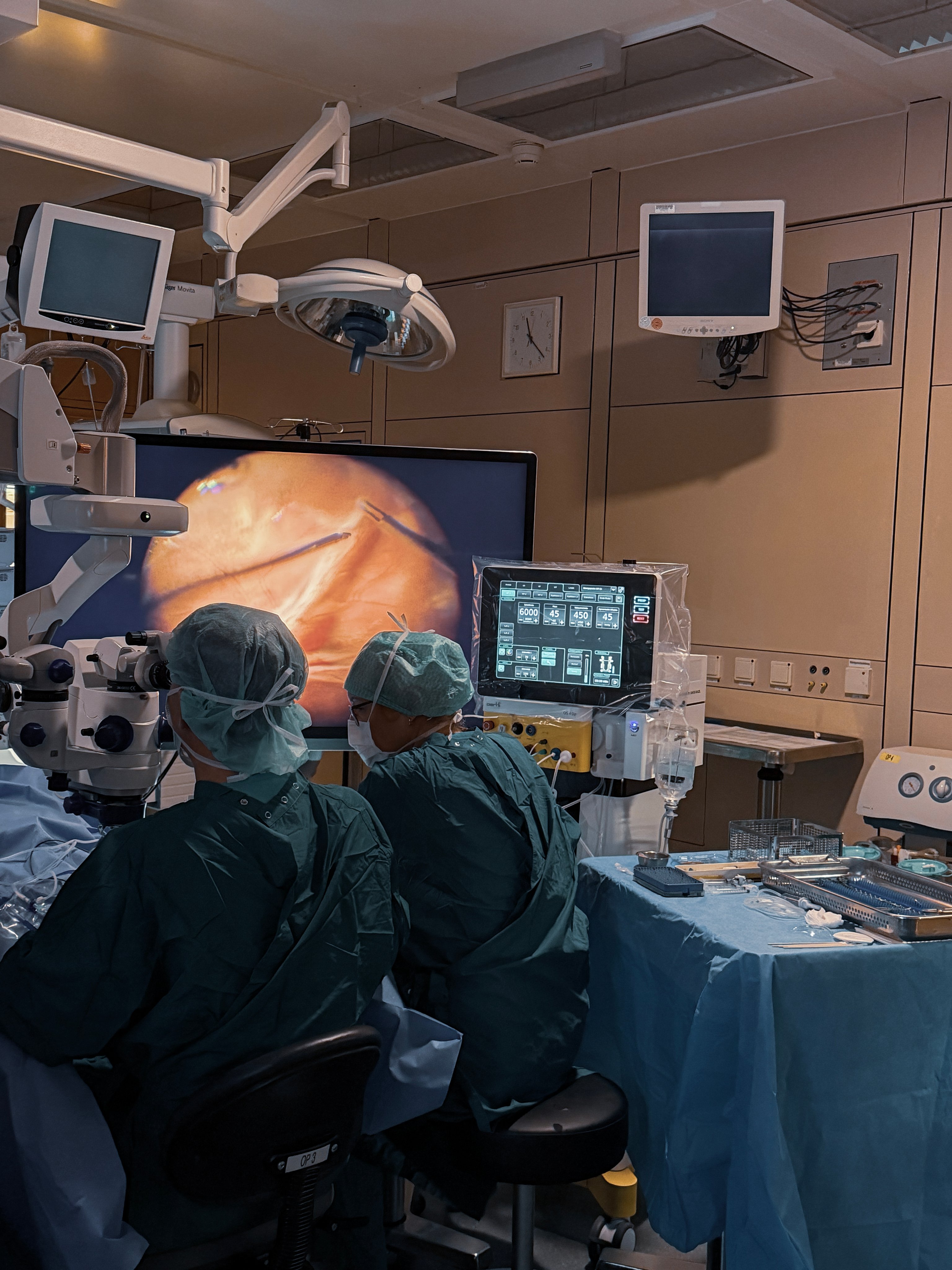 Operating room scene with two surgeons performing ophthalmic surgery under a microscope, supported by surgical platform and large display showing intraocular view