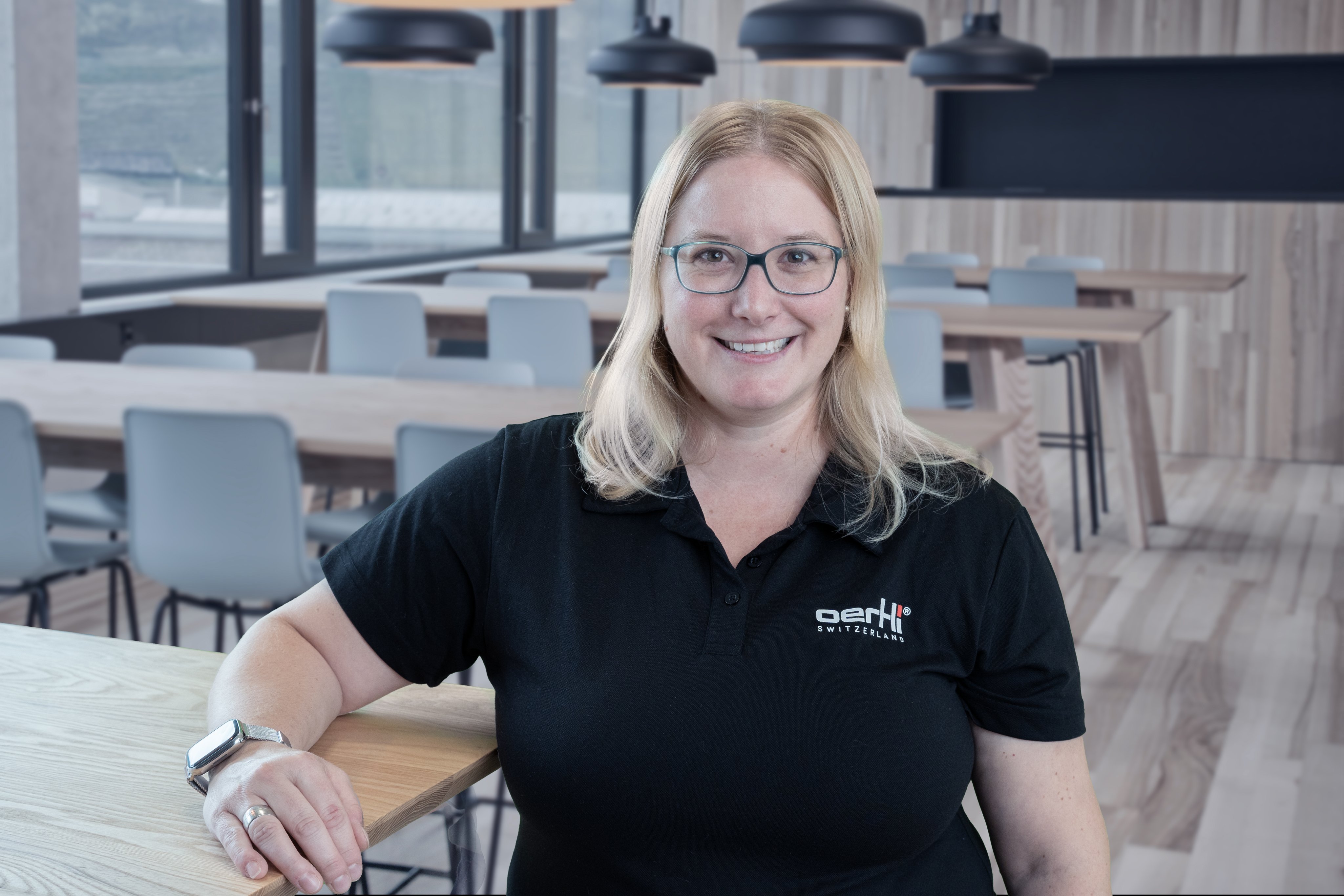 Portrait of a smiling Oertli employee wearing a black Oertli shirt in a modern cafeteria setting