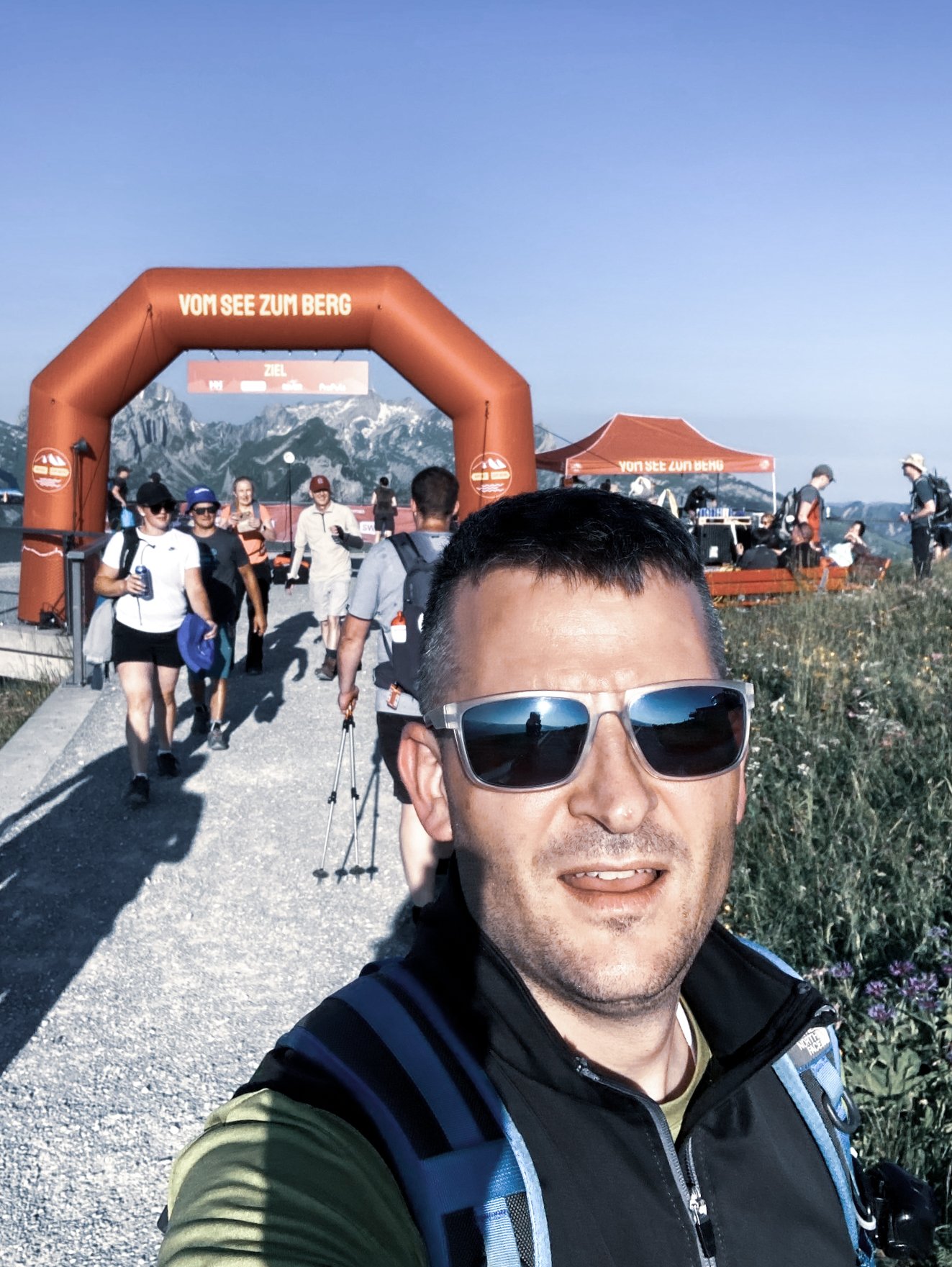 Mitarbeiter macht ein Selfie vor dem Zielbogen der Veranstaltung „Vom See zum Berg“ mit Bergkulisse im Hintergrund