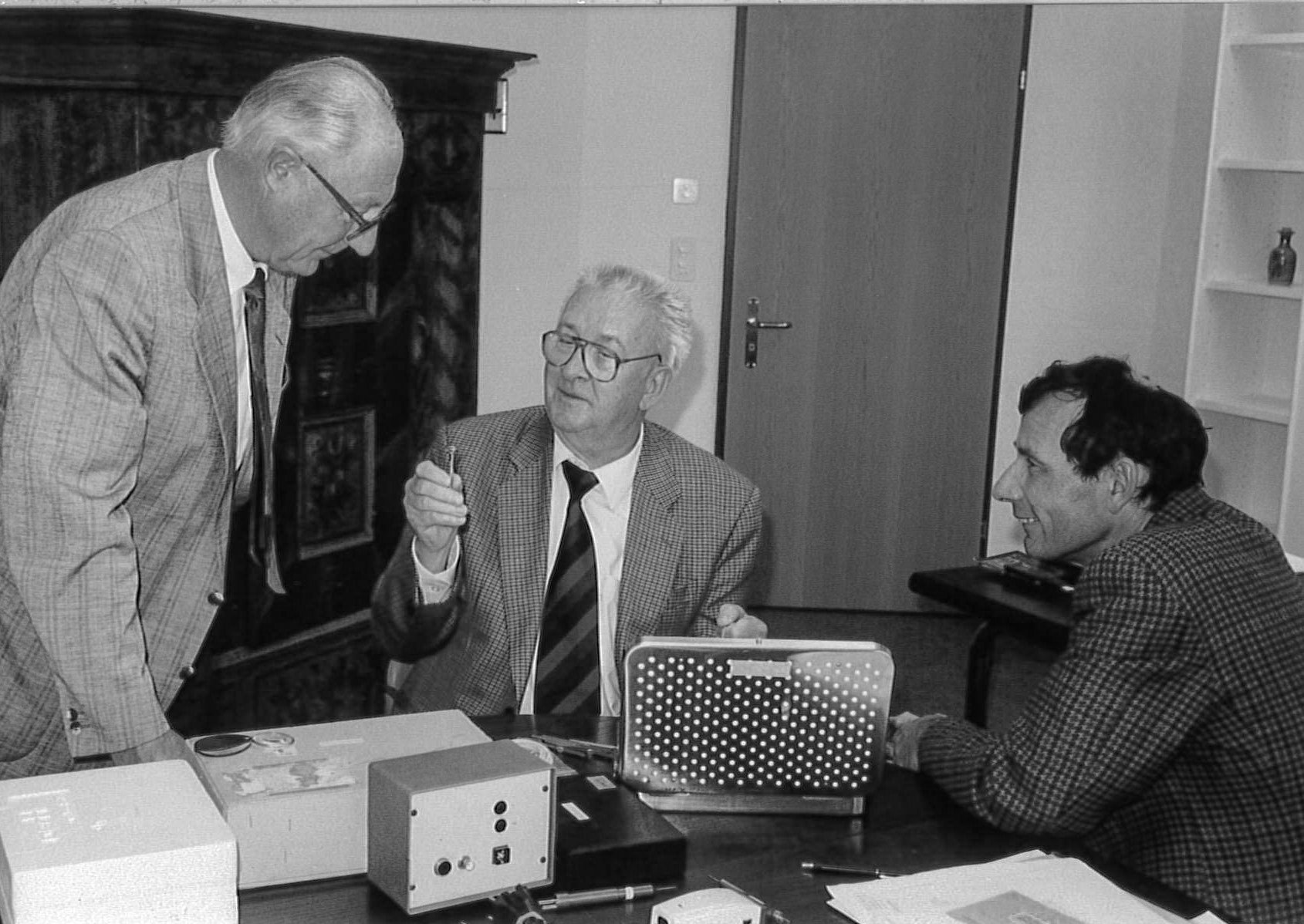 Black-and-white image of three men discussing an ophthalmic device and documents during a historical customer meeting