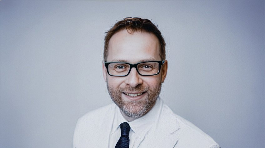 Portrait of male ophthalmologist wearing glasses and white coat, smiling against a neutral background