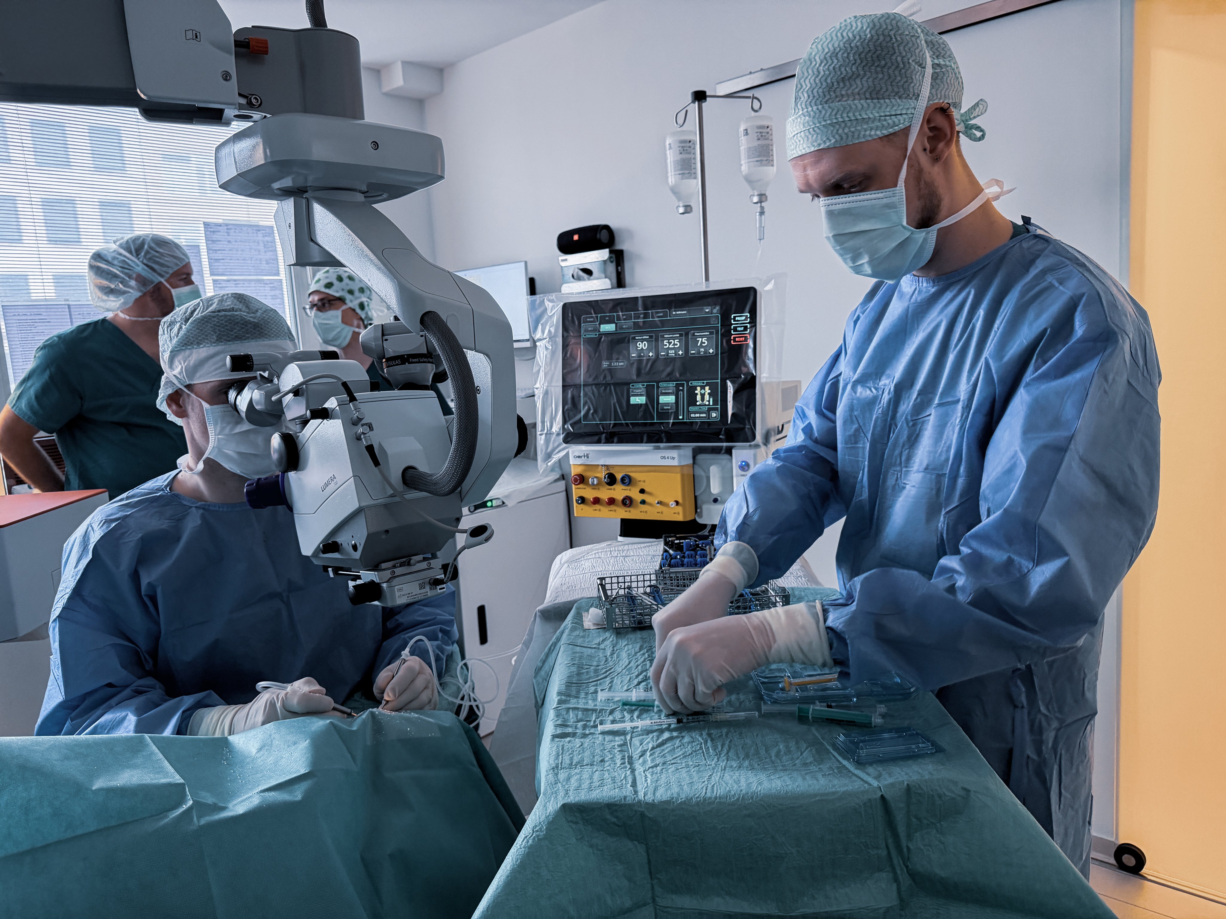 Ophthalmic surgery in operating room with surgeon using microscope and assistant preparing instruments beside surgical system OS 4 Up