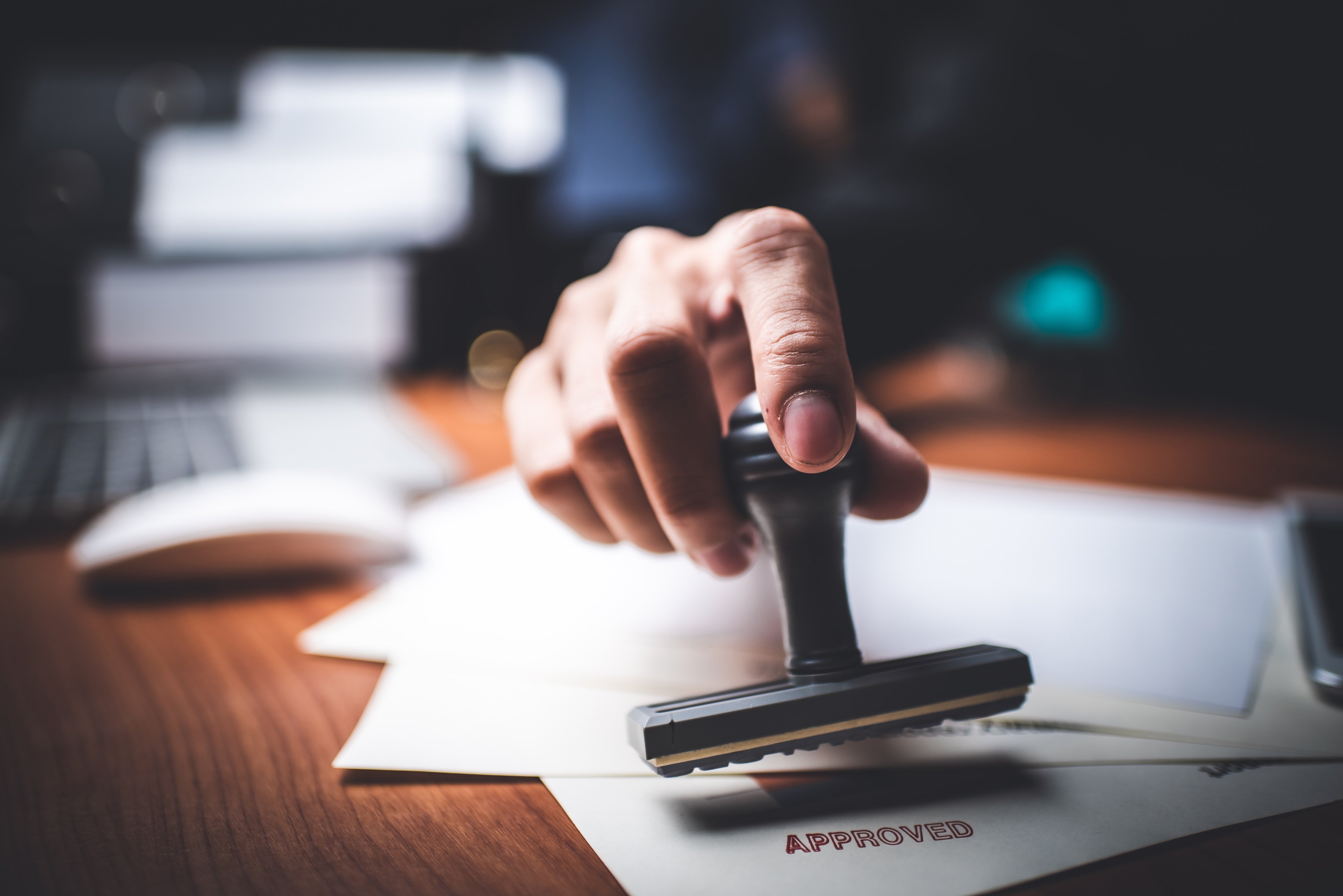 Hand using a stamp on official documents beside a laptop and office desk, symbolising regulatory approval and medical compliance procedures.