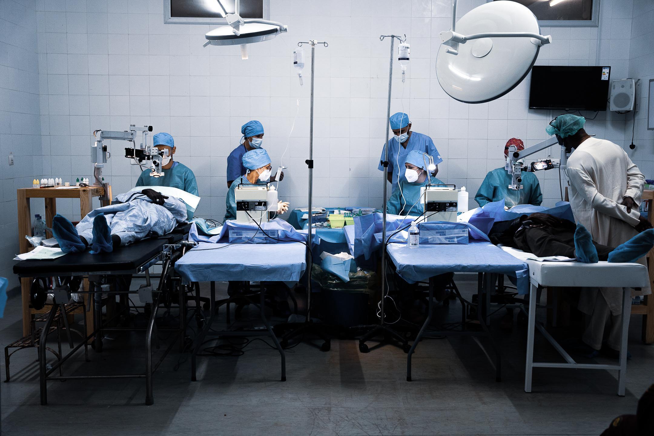 Wide view of an ophthalmic operating room with multiple surgical teams performing eye surgery during a charitable medical outreach mission.