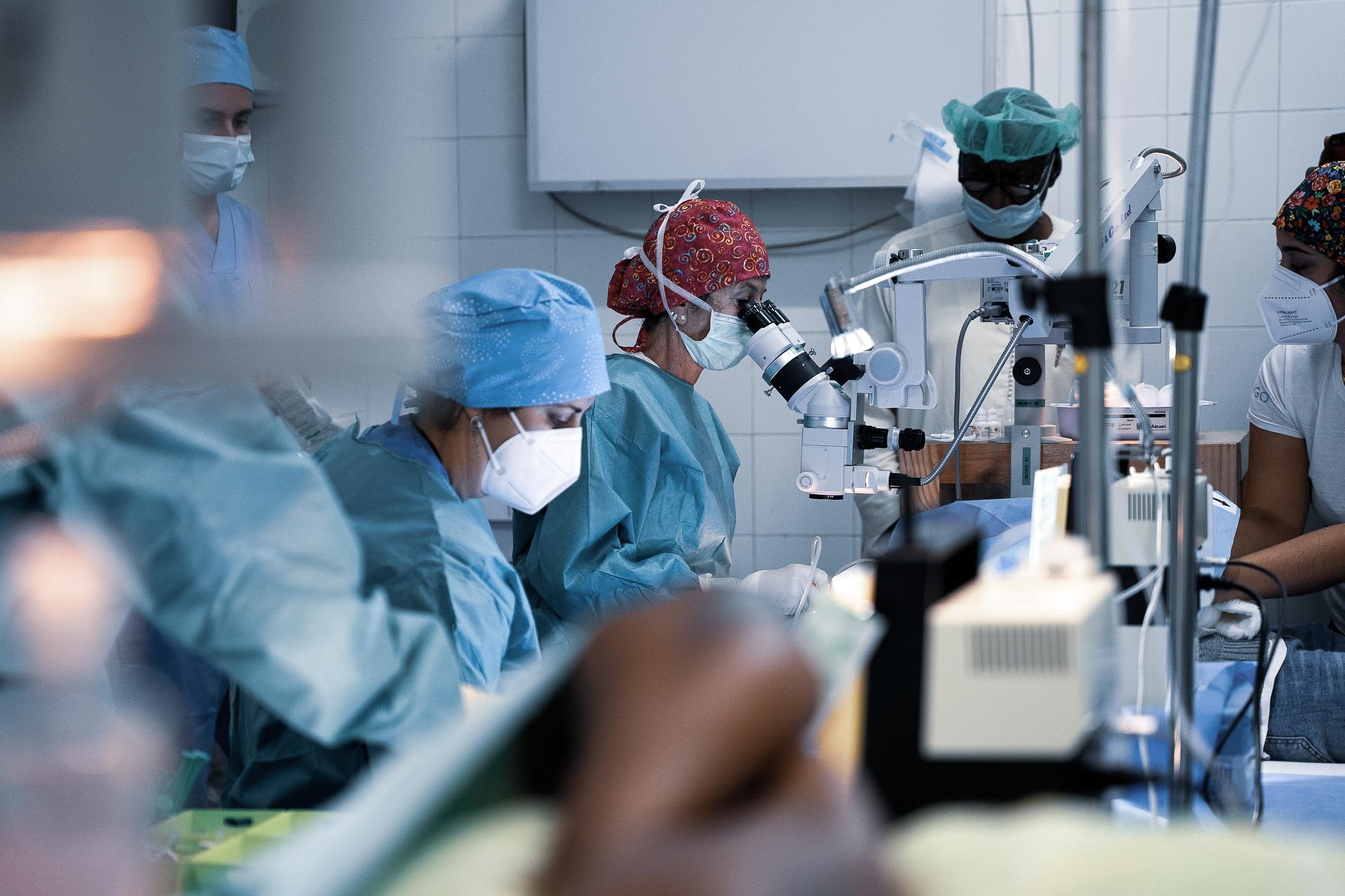 Medical team performing eye surgery in an operating room, with surgeons wearing masks and surgical caps while working at ophthalmic microscopes and surgical systems.