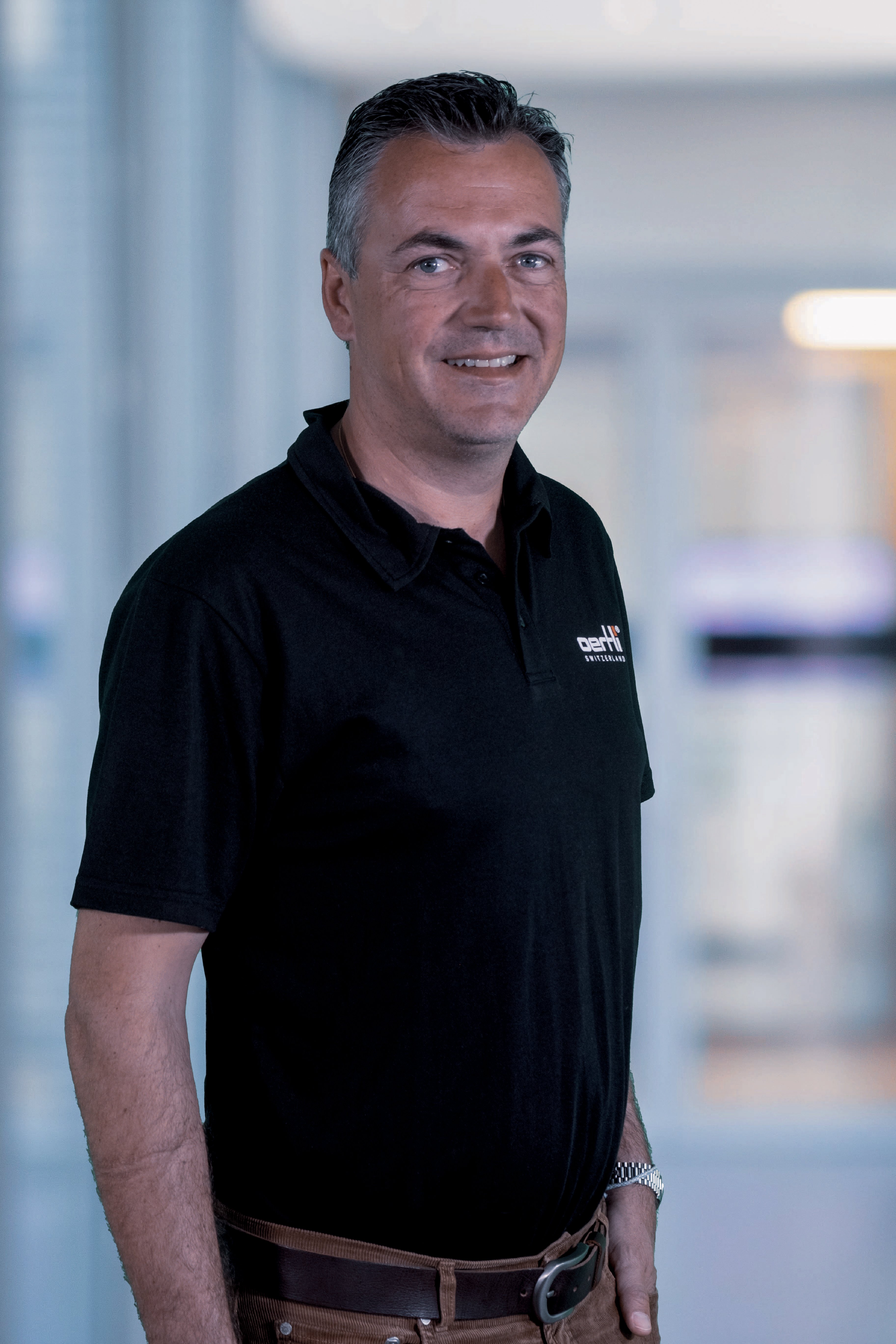 Portrait of a smiling Oertli employee wearing a black Oertli shirt against a softly blurred office background