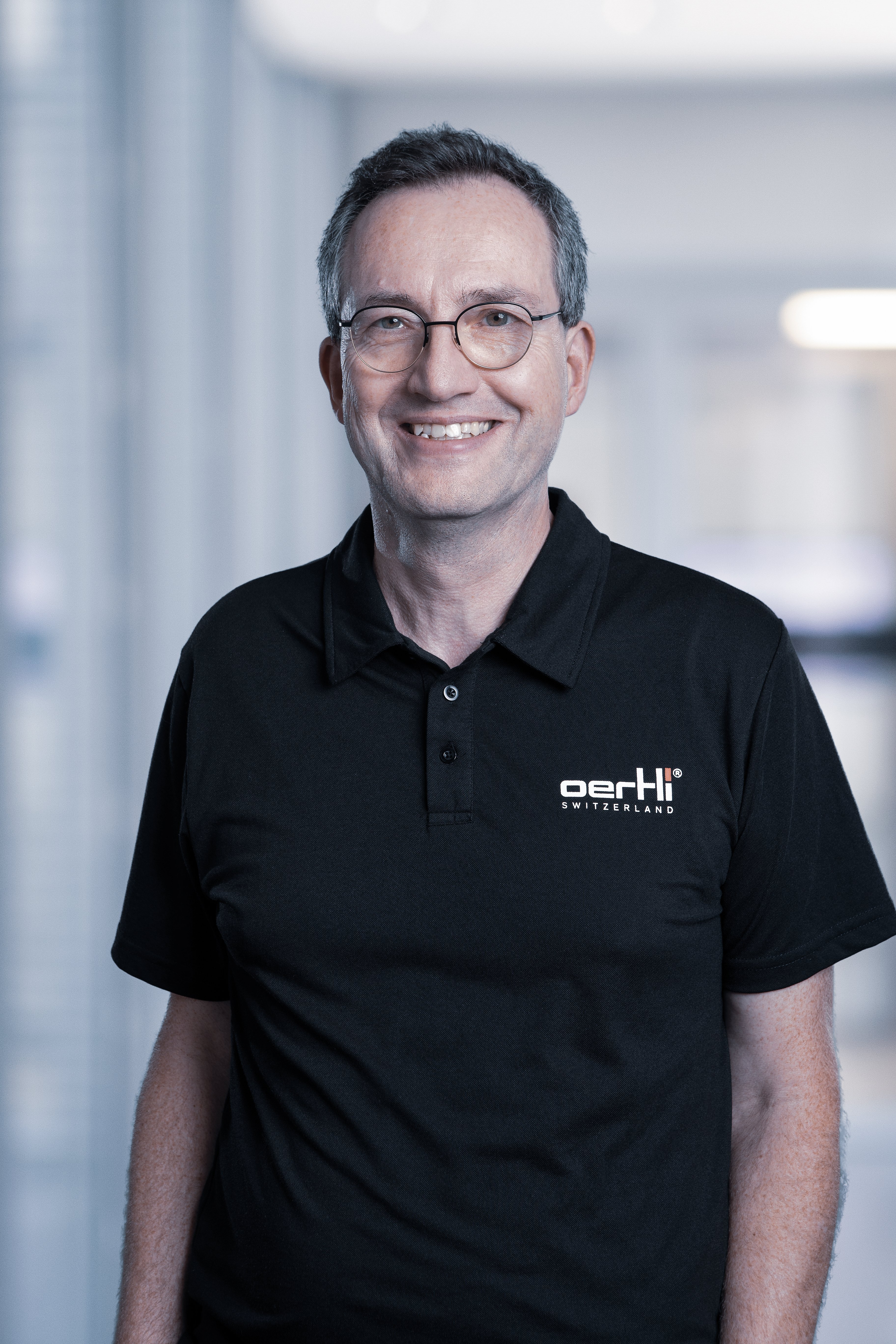 Portrait of a smiling Oertli employee wearing glasses and a black Oertli shirt against a softly blurred office background