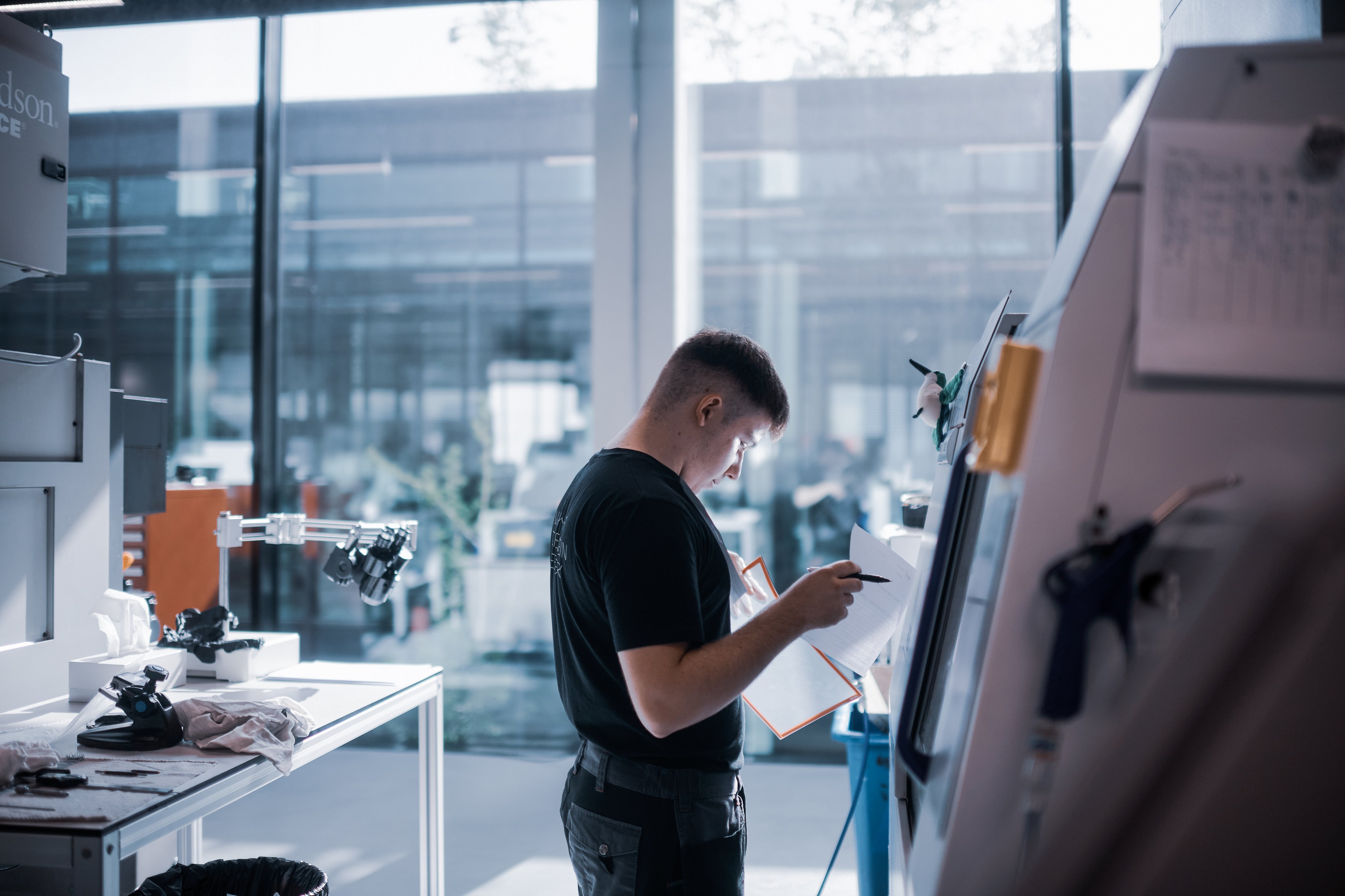 Lernender arbeitet in der Teilefertigung an einer Fertigungsmaschine und prüft Produktionsunterlagen in einer modernen Produktionsumgebung