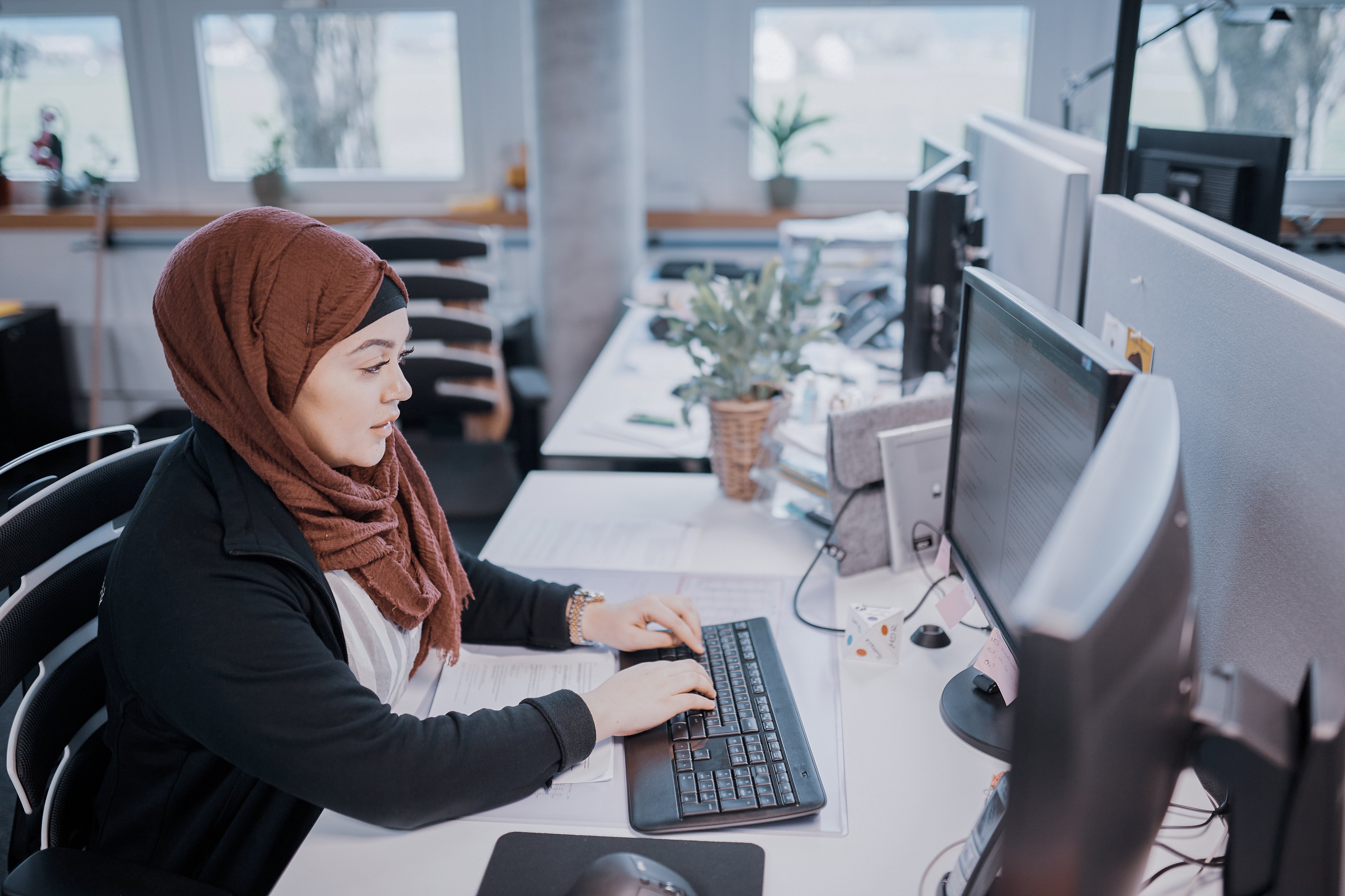 Woman working at a desk with multiple computer monitors in a bright office environment, focused on digital and administrative tasks.