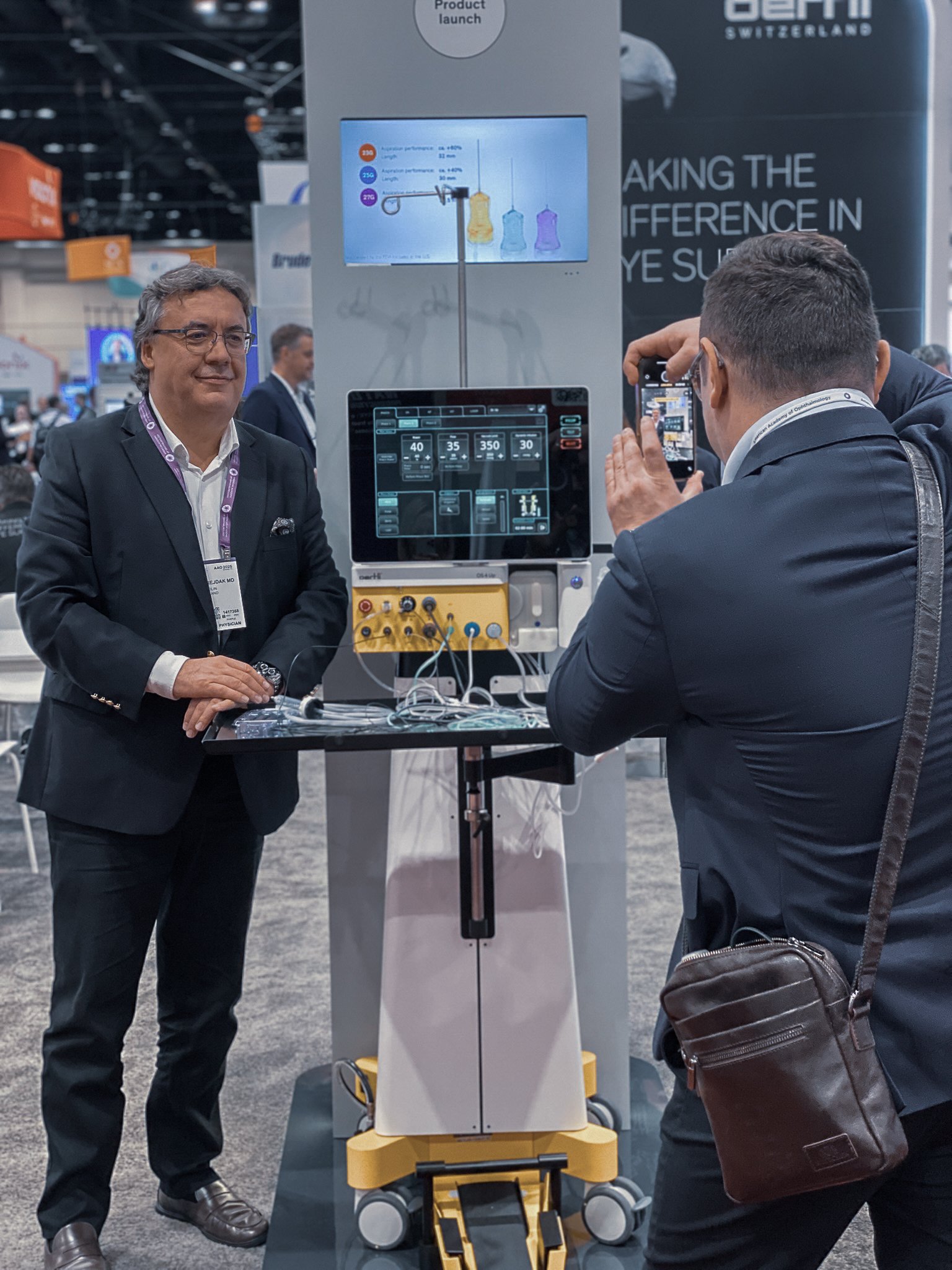 A visitor standing beside the Oertli surgical platform OS 4 Up at AAO 2025 while another attendee takes a photograph. The device is displayed at the Oertli exhibition booth during the ophthalmology congress