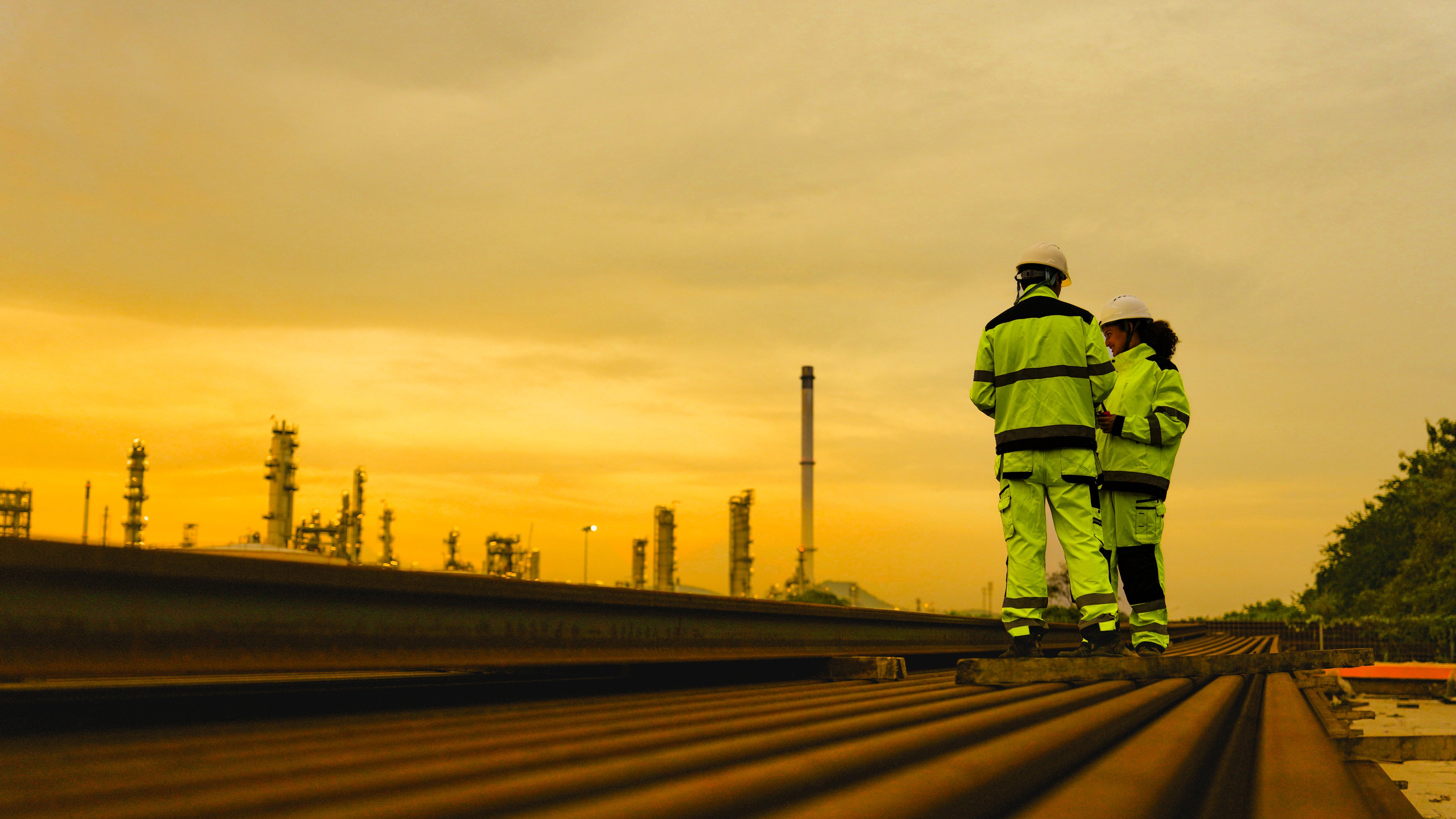 Two industrial workers in high visibility safety gear and hard hats stand on rusted metal pipes. They are inspecting a large factory or refinery plant during a golden sunset.