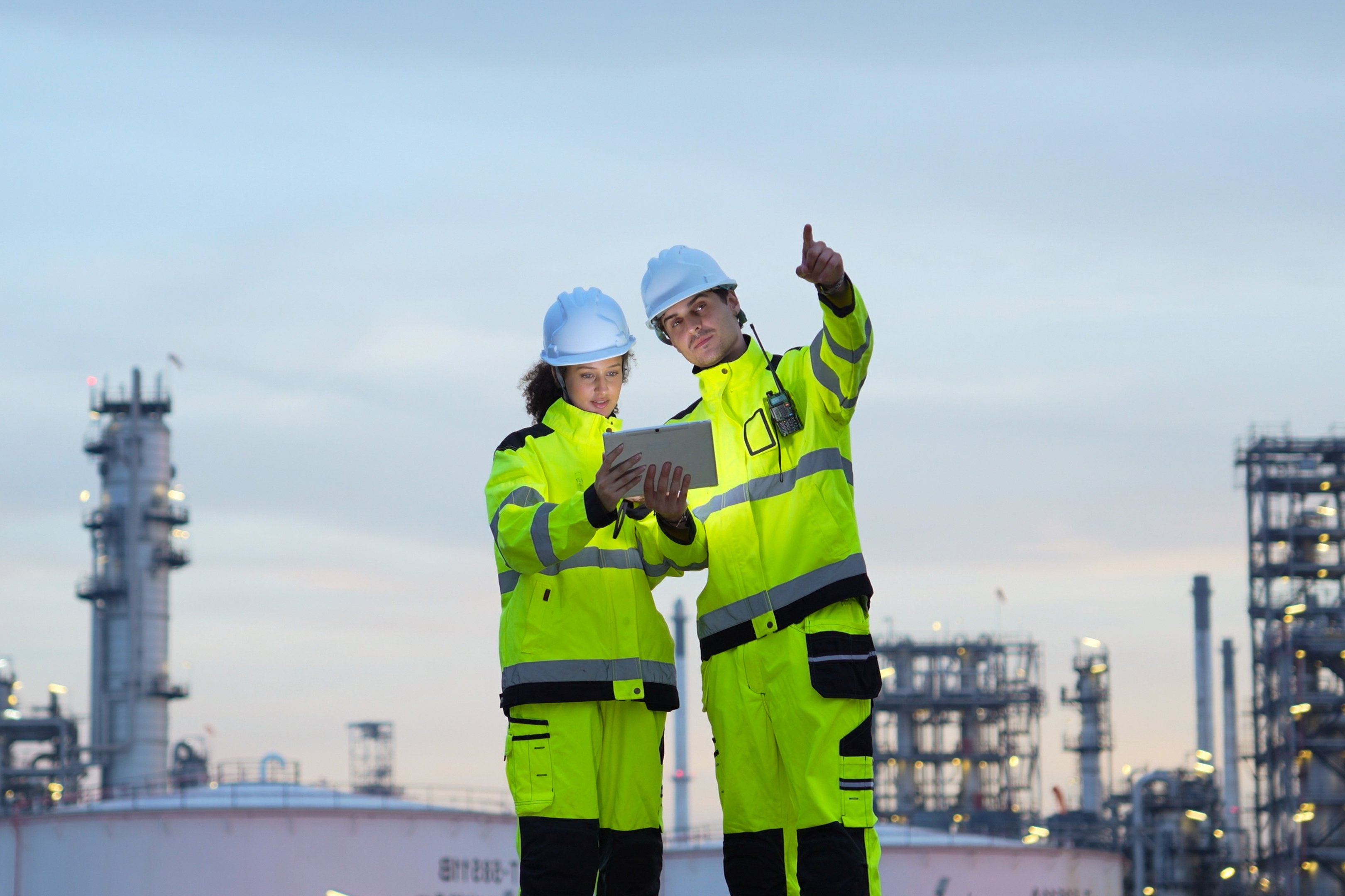 Safety Engineers Surveying Refinery Operations, Engineers Reviewing Plans at Industrial Site at Dusk, Team of Industrial Workers with Tablet at Night